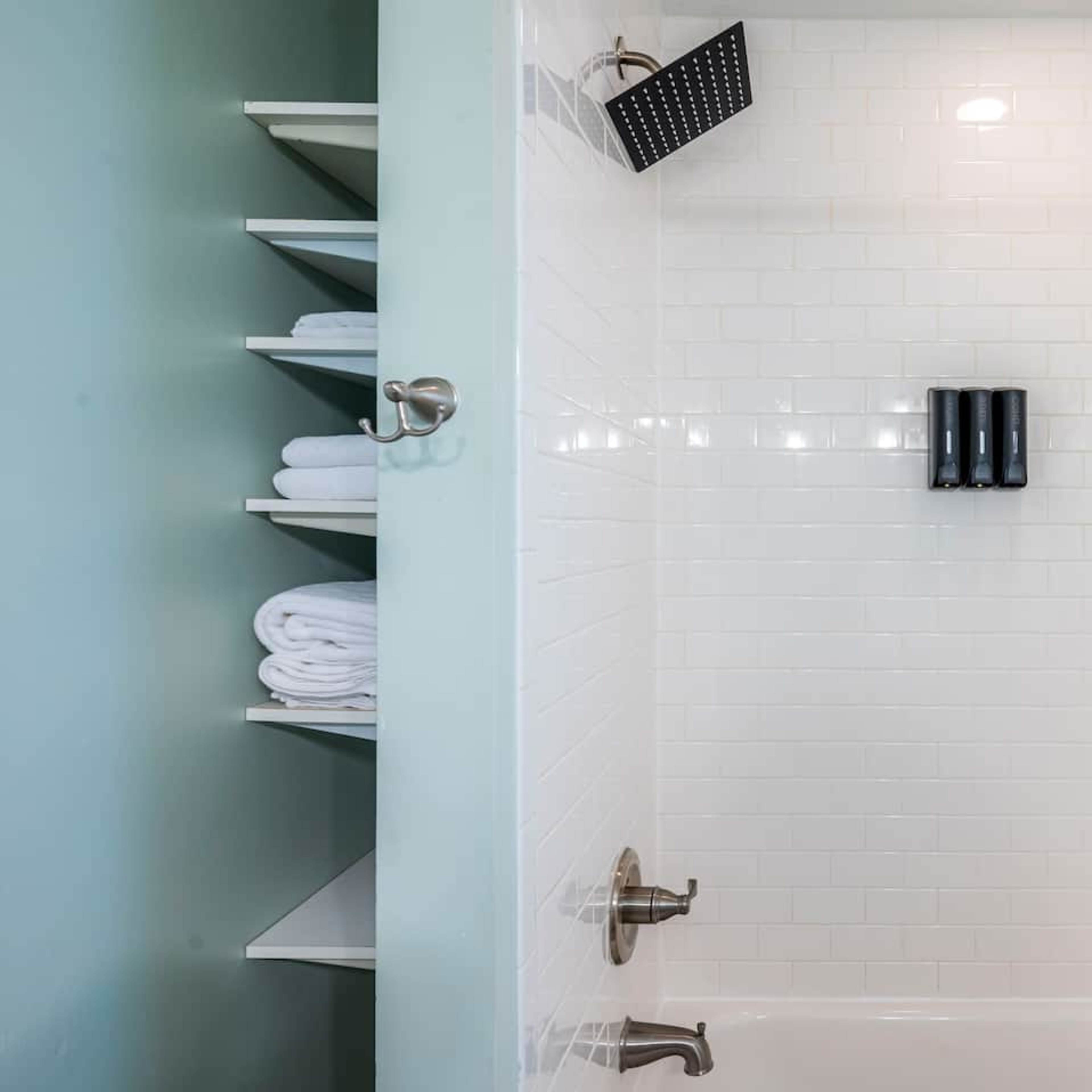 A clean shower area with white tile walls, a showerhead, and neatly arranged towels on shelves.