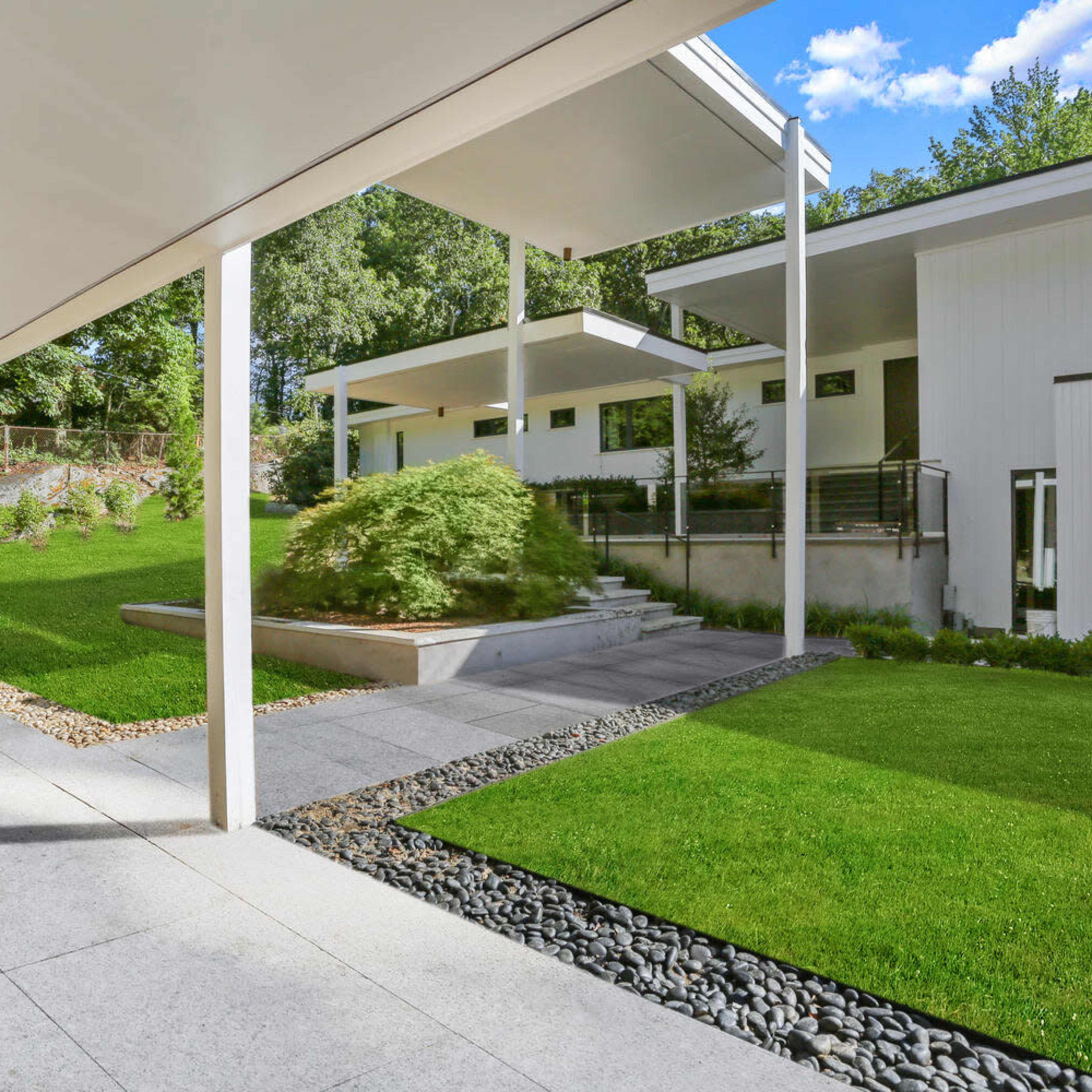 A modern house is set on a landscaped yard with manicured grass and stone pathways leading to a raised patio.