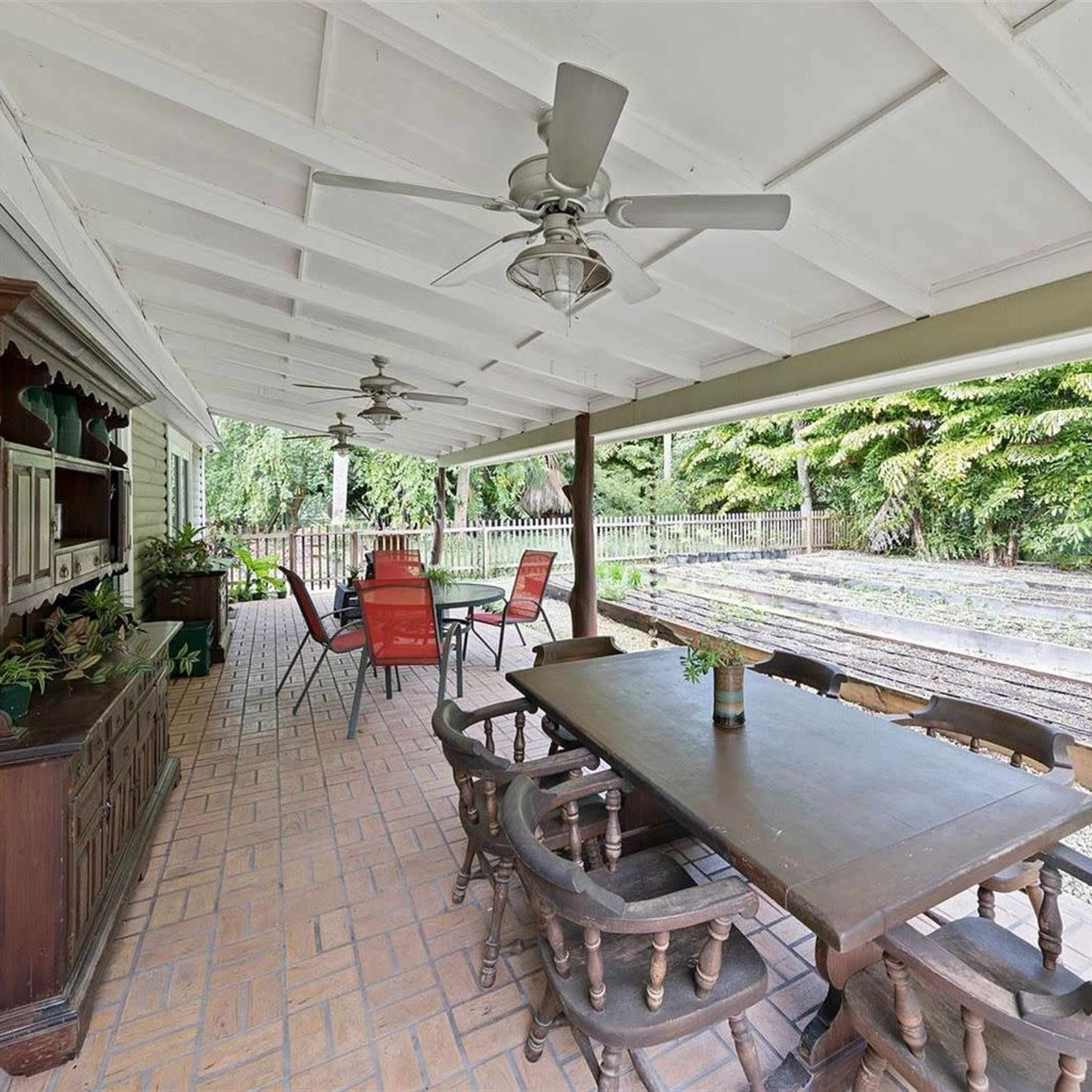 A covered patio with a wooden dining table, chairs, and ceiling fans, overlooking a fenced garden area with raised planting beds.