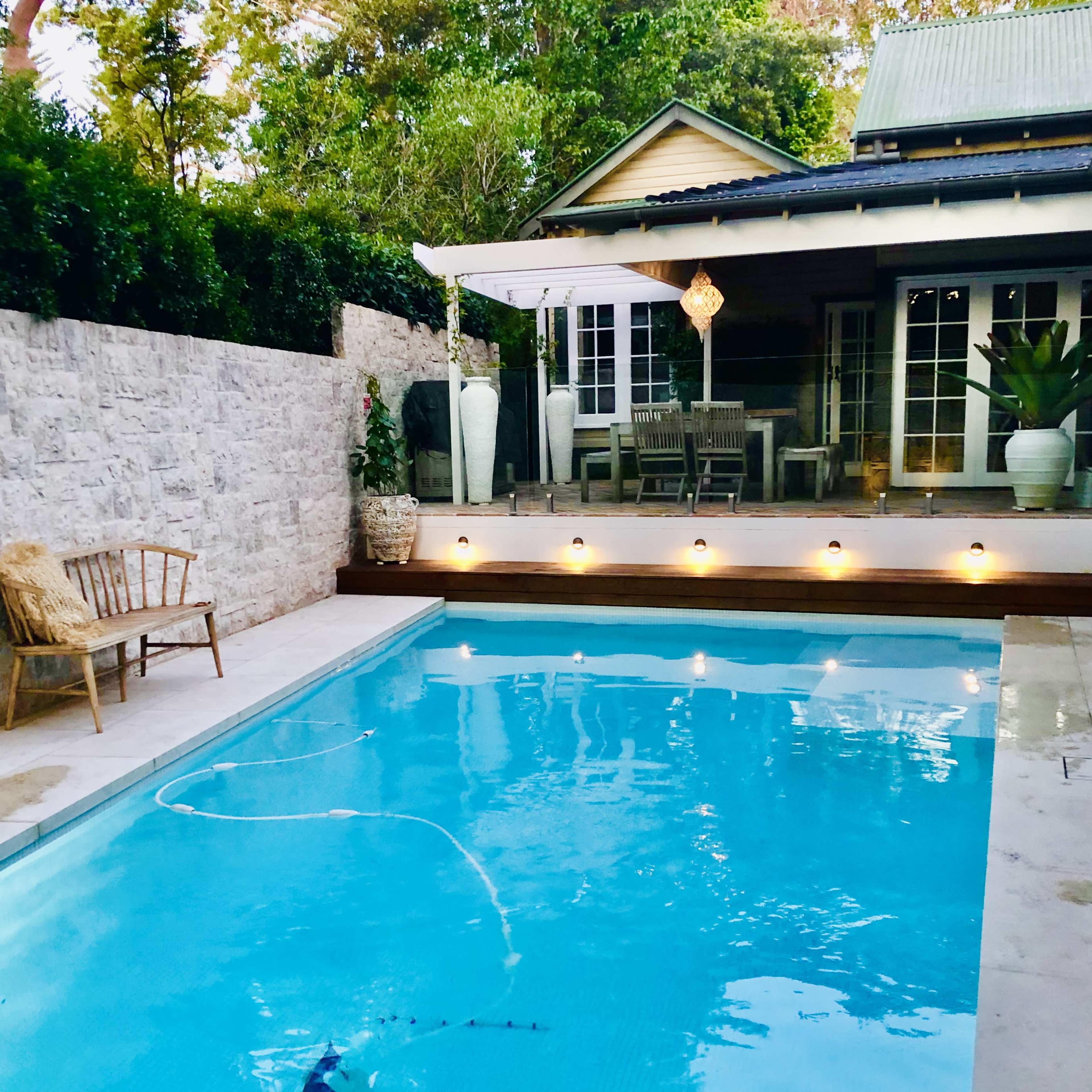 A backyard with a swimming pool, surrounded by a stone wall, patio furniture, and a house with a deck.