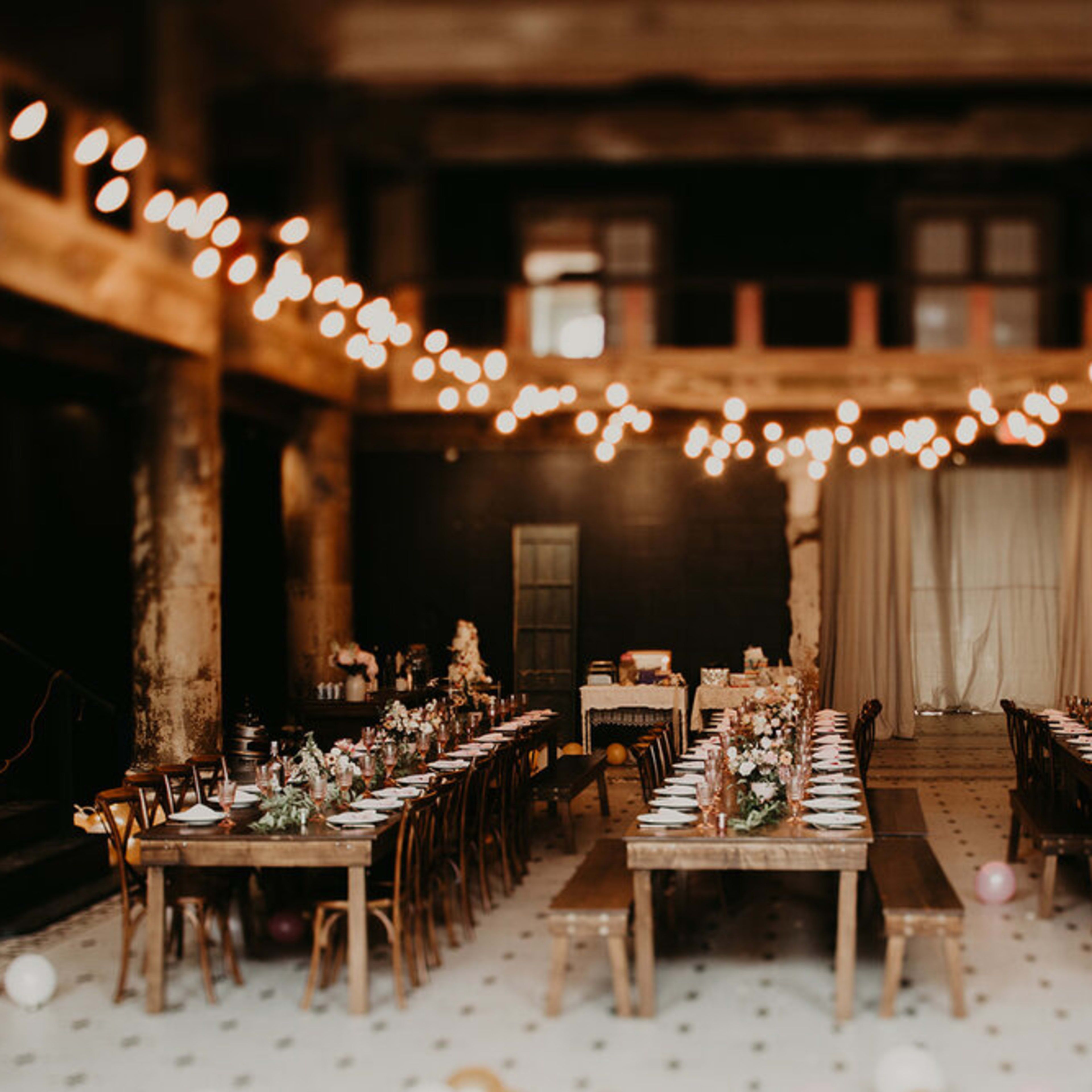 A long banquet hall features multiple wooden tables set for dining, adorned with floral centerpieces and string lights overhead.