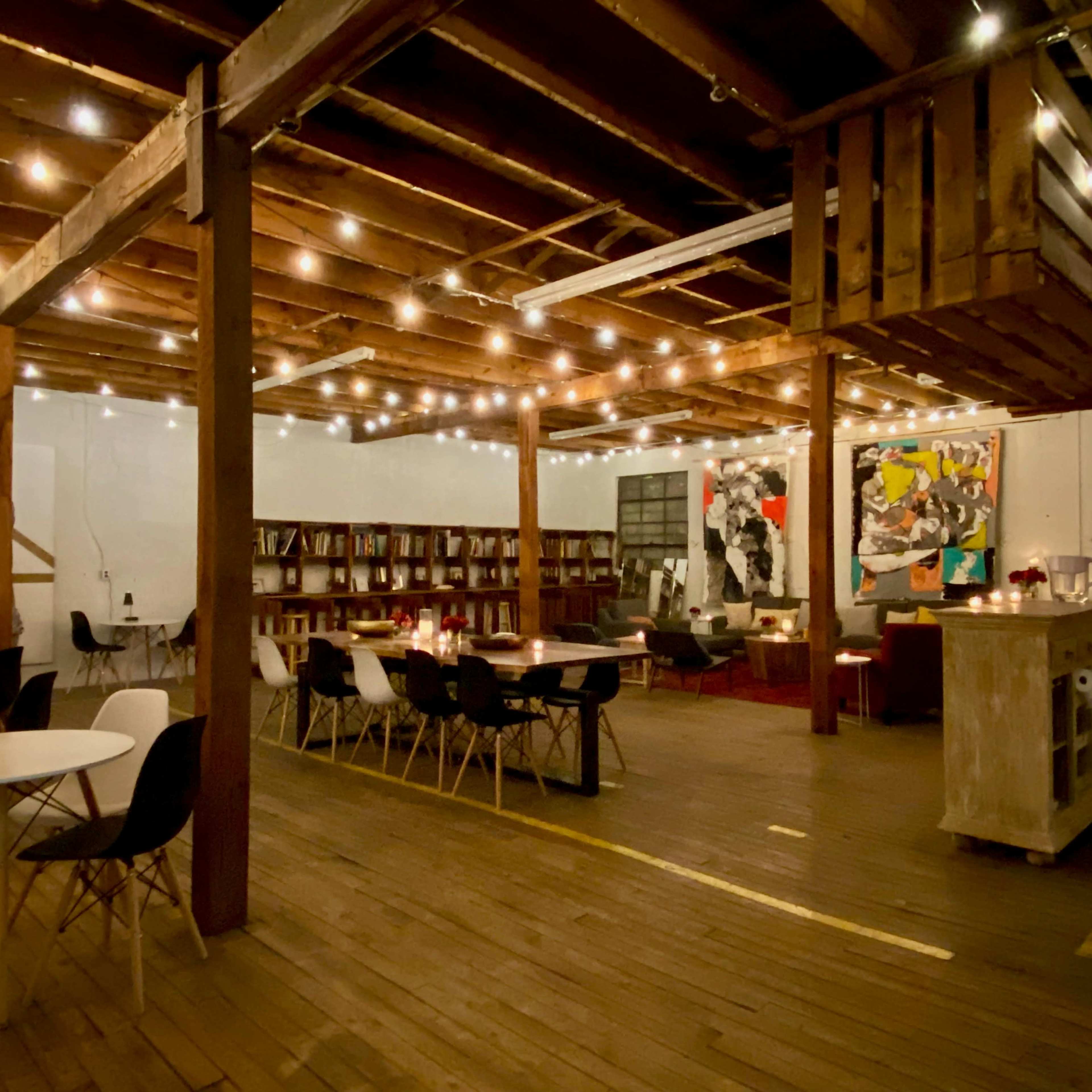 A spacious interior with wooden beams, string lights, a collection of bookshelves, and various seating arrangements, including tables and modern chairs.