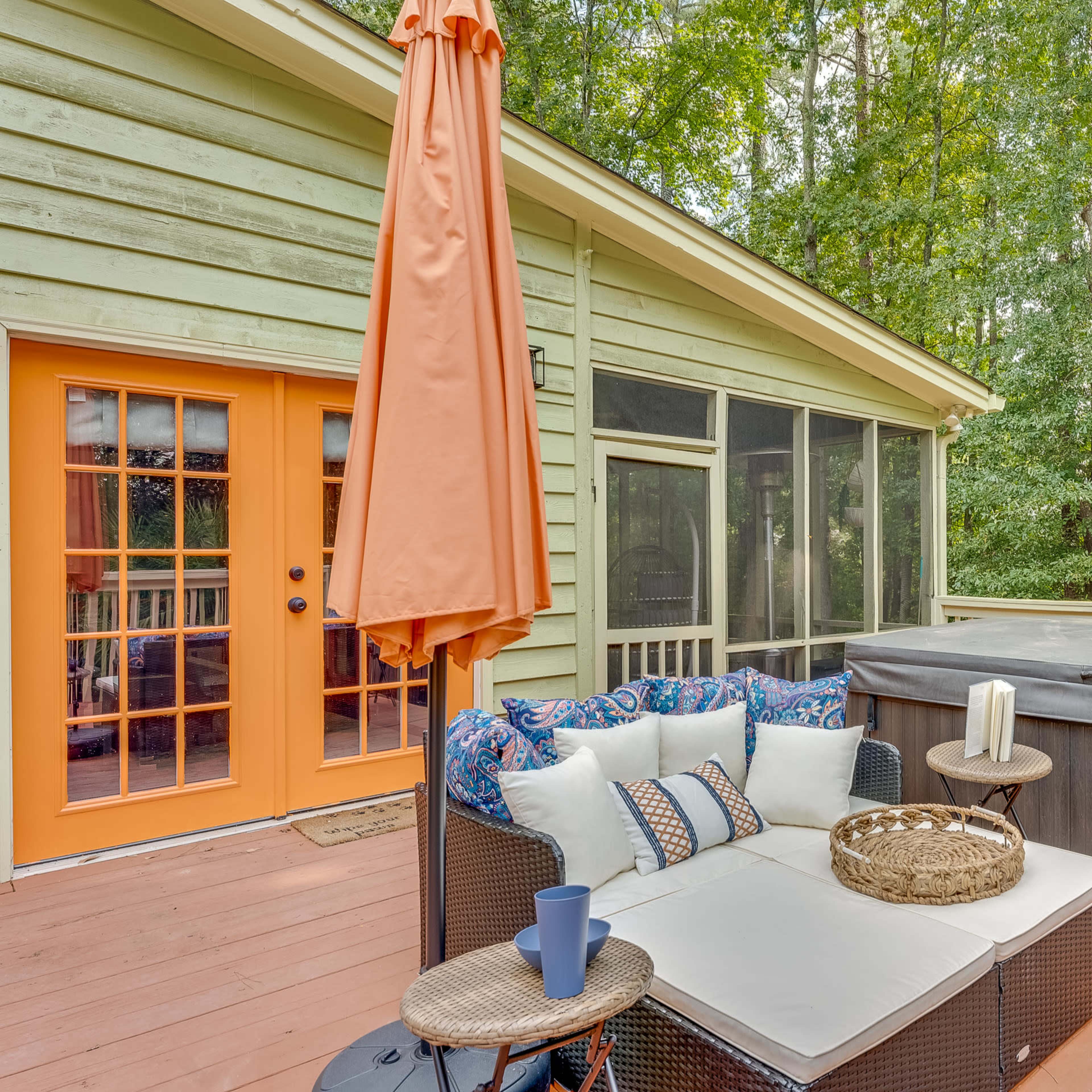 A wooden deck features a hot tub, a cozy seating area with an umbrella, and a backdrop of tall green trees.