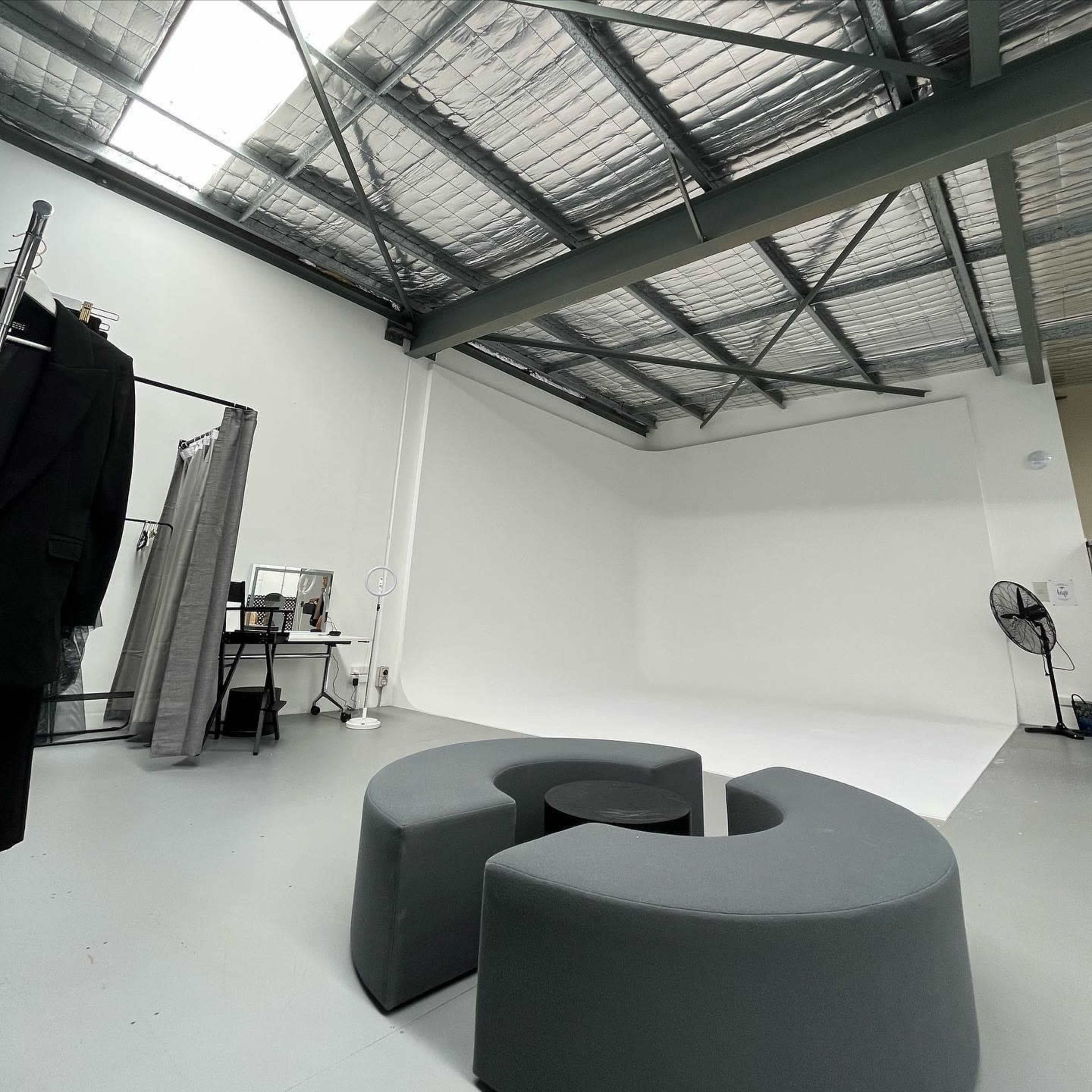 A minimalist studio space with curved gray seating, a black table, and various props including a mirror and a clothing rack against white walls.