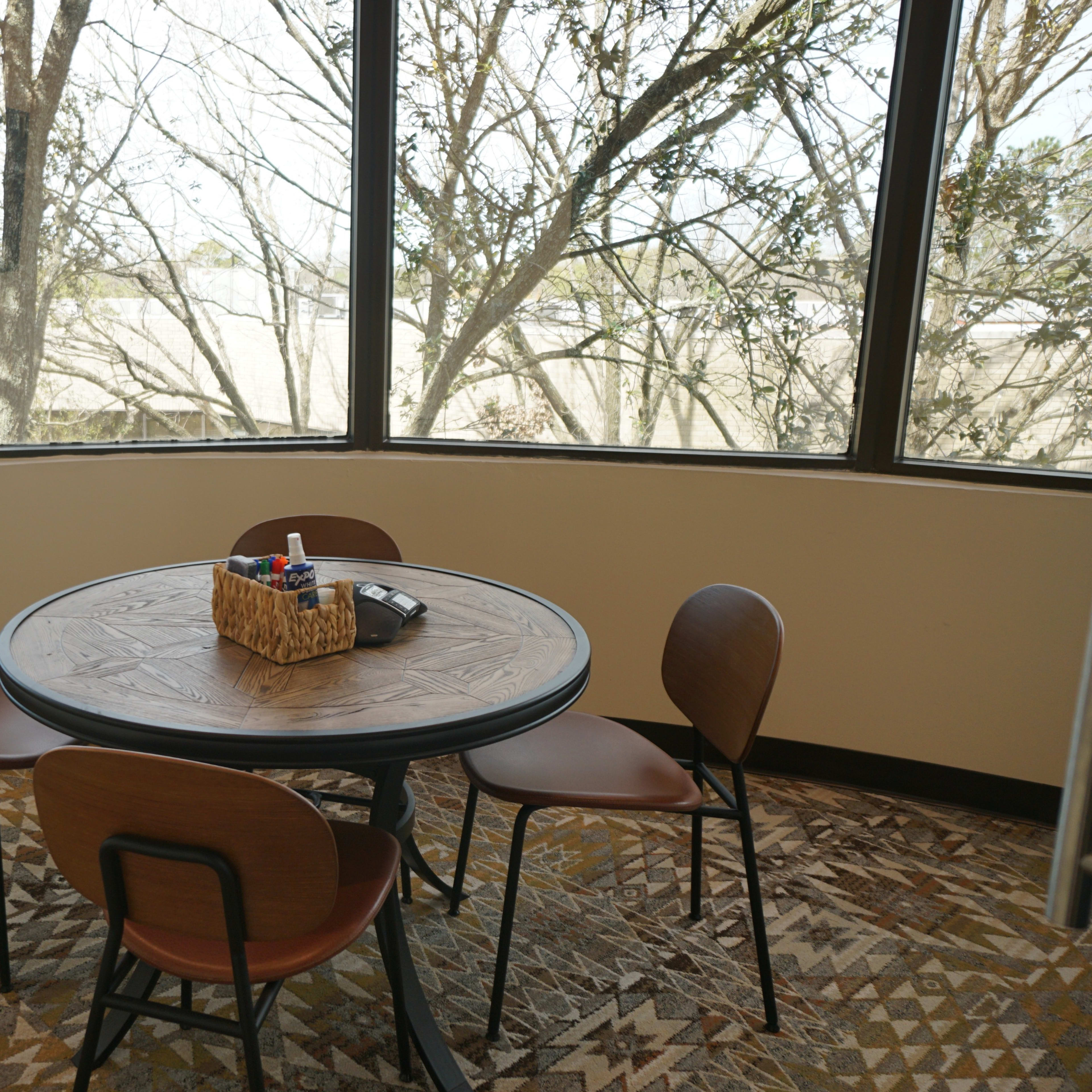 A round table with a basket of markers sits surrounded by four chairs in a room with large windows displaying trees outside.