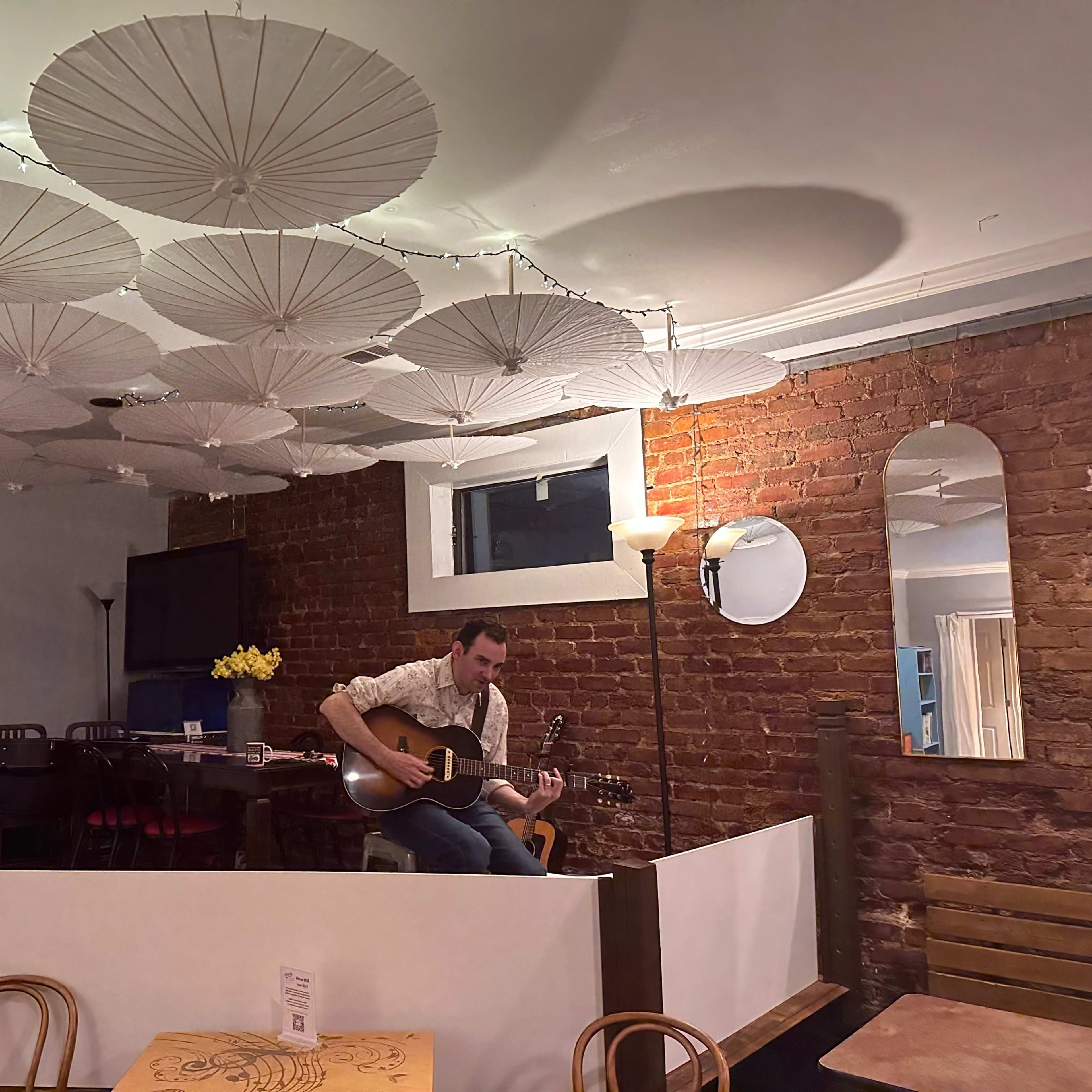 A musician sits on a stool playing guitar in a cozy cafe with a brick wall and paper umbrellas hanging from the ceiling.