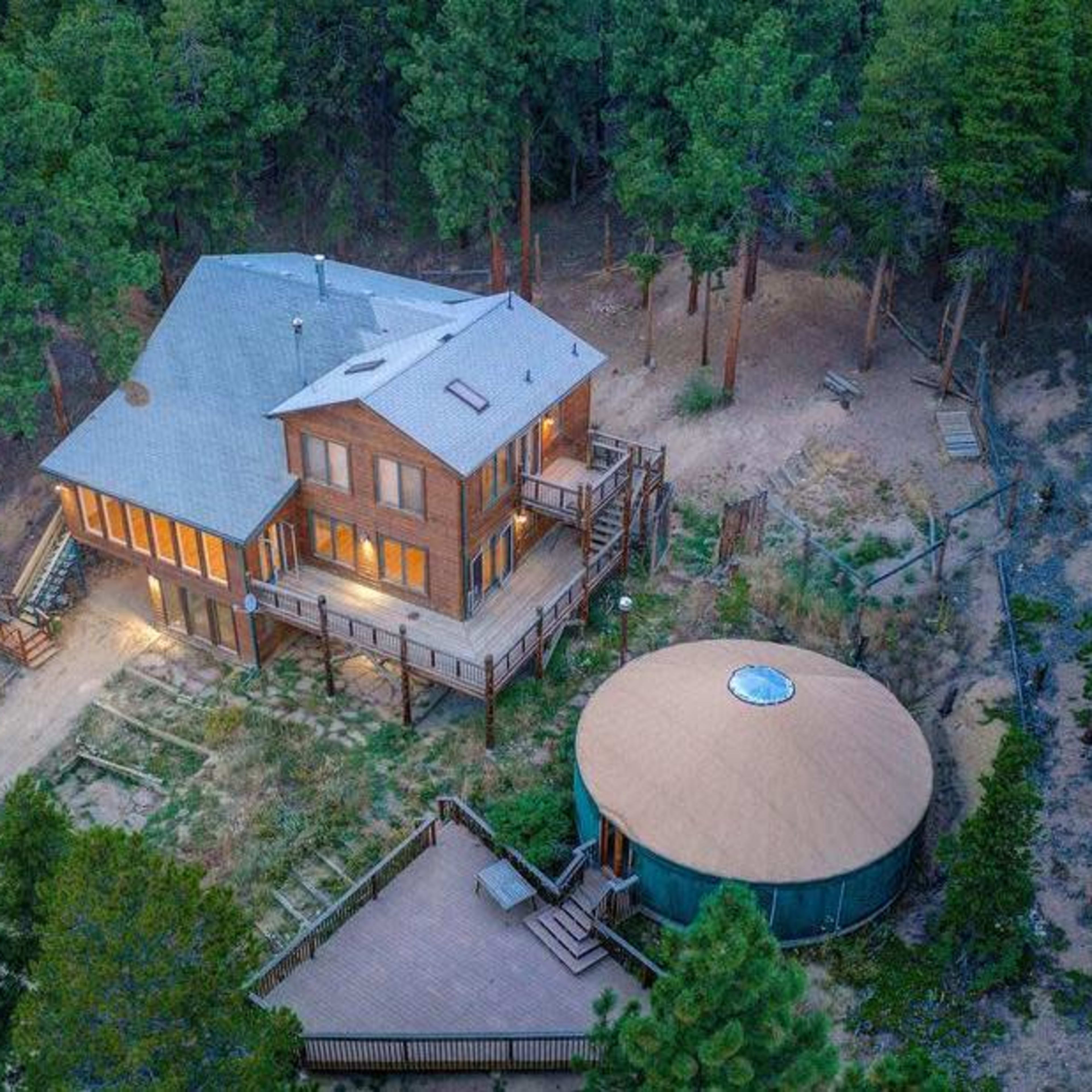 A large wooden house with multiple levels and large windows is surrounded by tall trees, with a circular structure and wooden deck nearby.