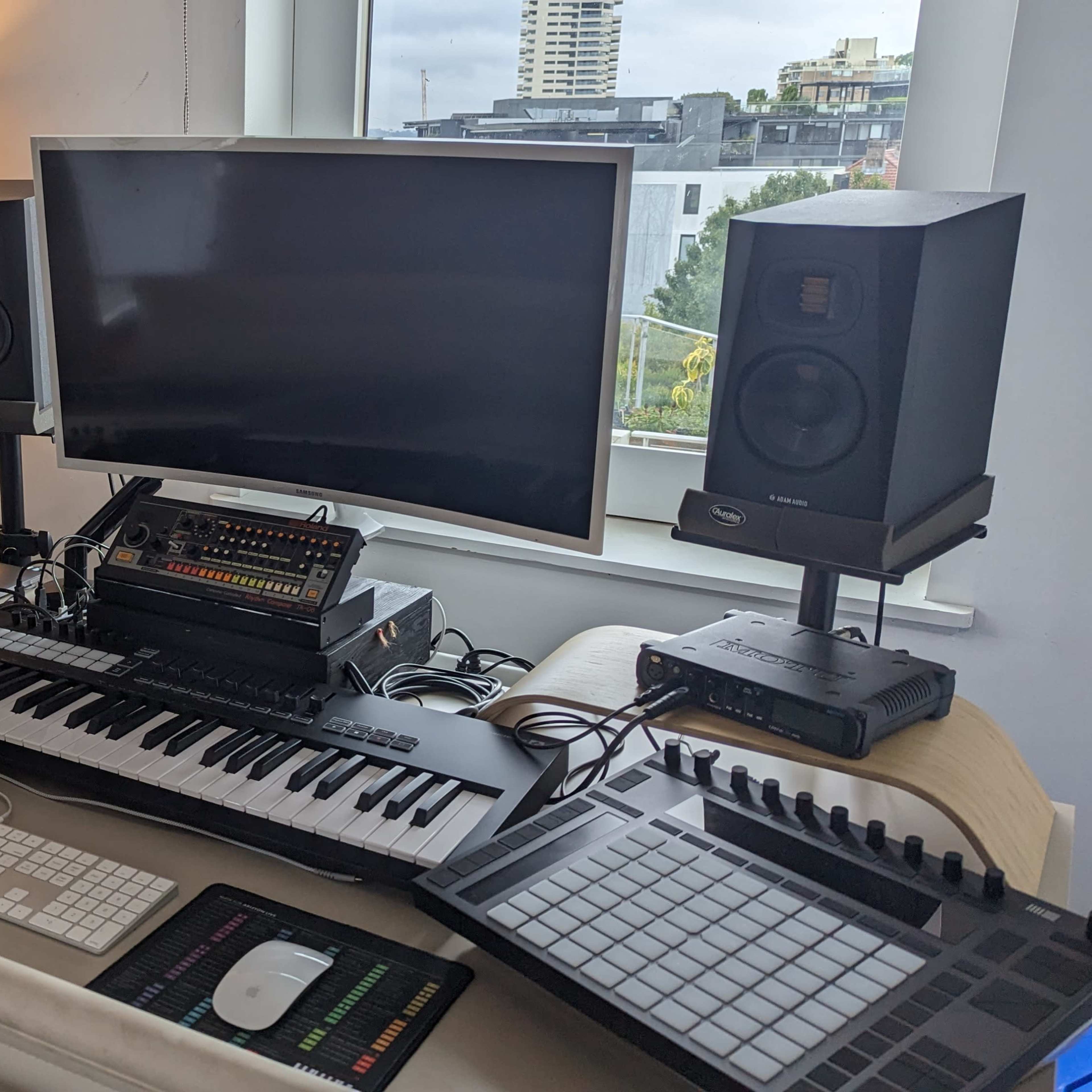 A modern music production workspace featuring a computer monitor, audio speakers, a keyboard, and various electronic instruments, with guitars positioned in the background.