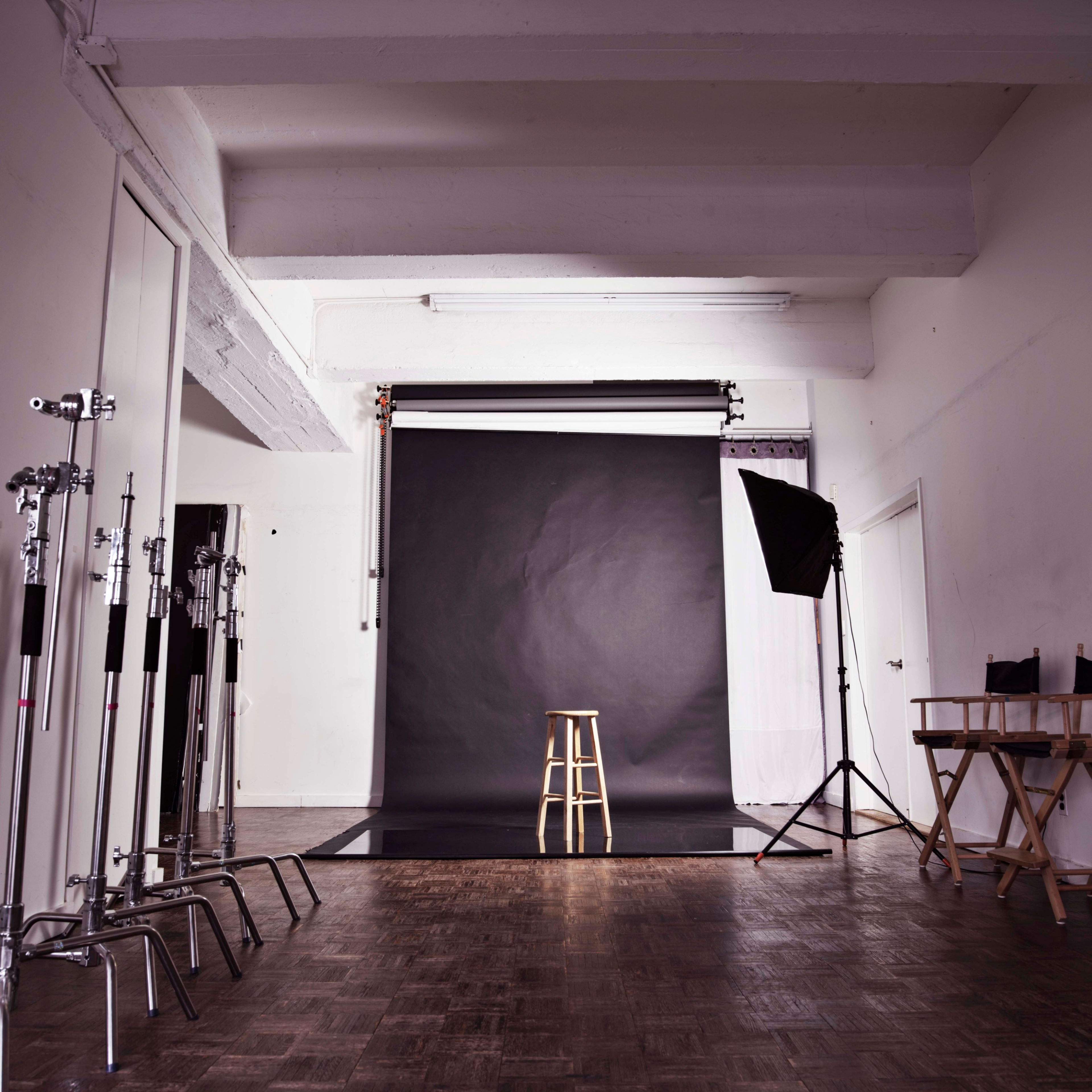 A photography studio features a black backdrop, a wooden stool in the center, and various lighting equipment along the sides.
