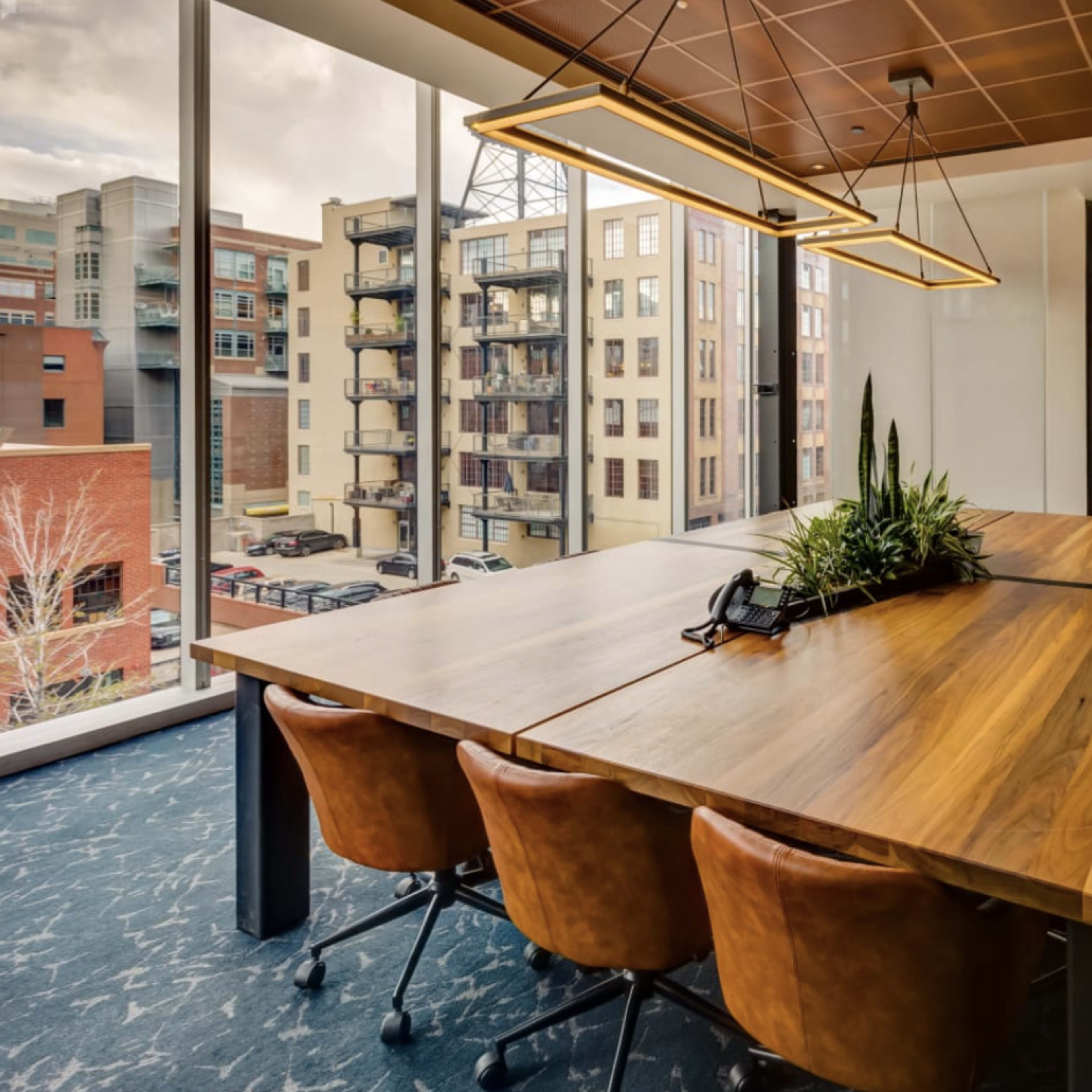 A large wooden conference table with brown chairs is situated in a well-lit office with large windows showing a view of neighboring buildings.