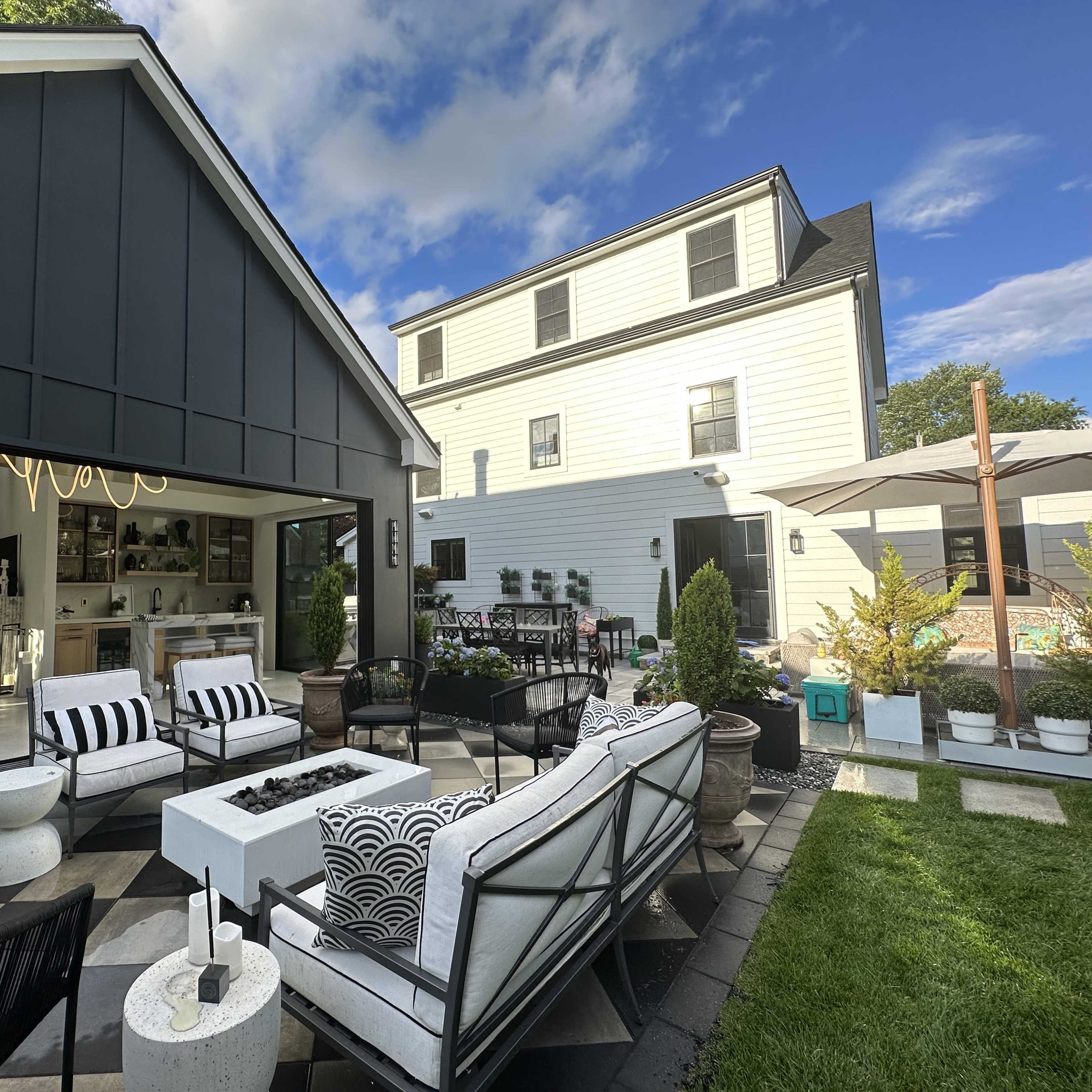 A modern outdoor seating area with a fire pit, surrounded by greenery, leading up to a white house and adjacent black-clad structure.