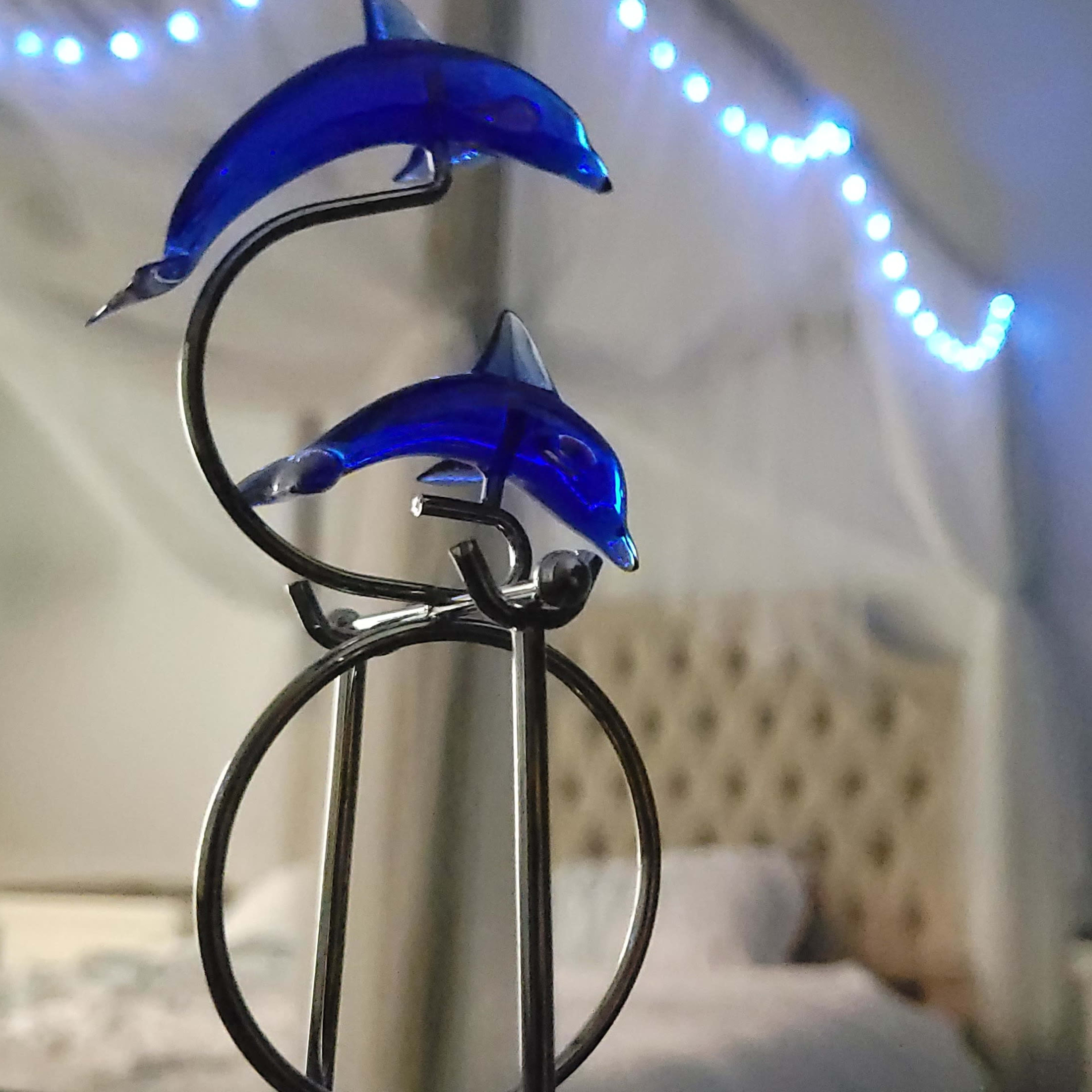 A metal holder featuring two blue dolphin figurines is placed in front of a softly lit bedroom with a bed and decorative string lights.