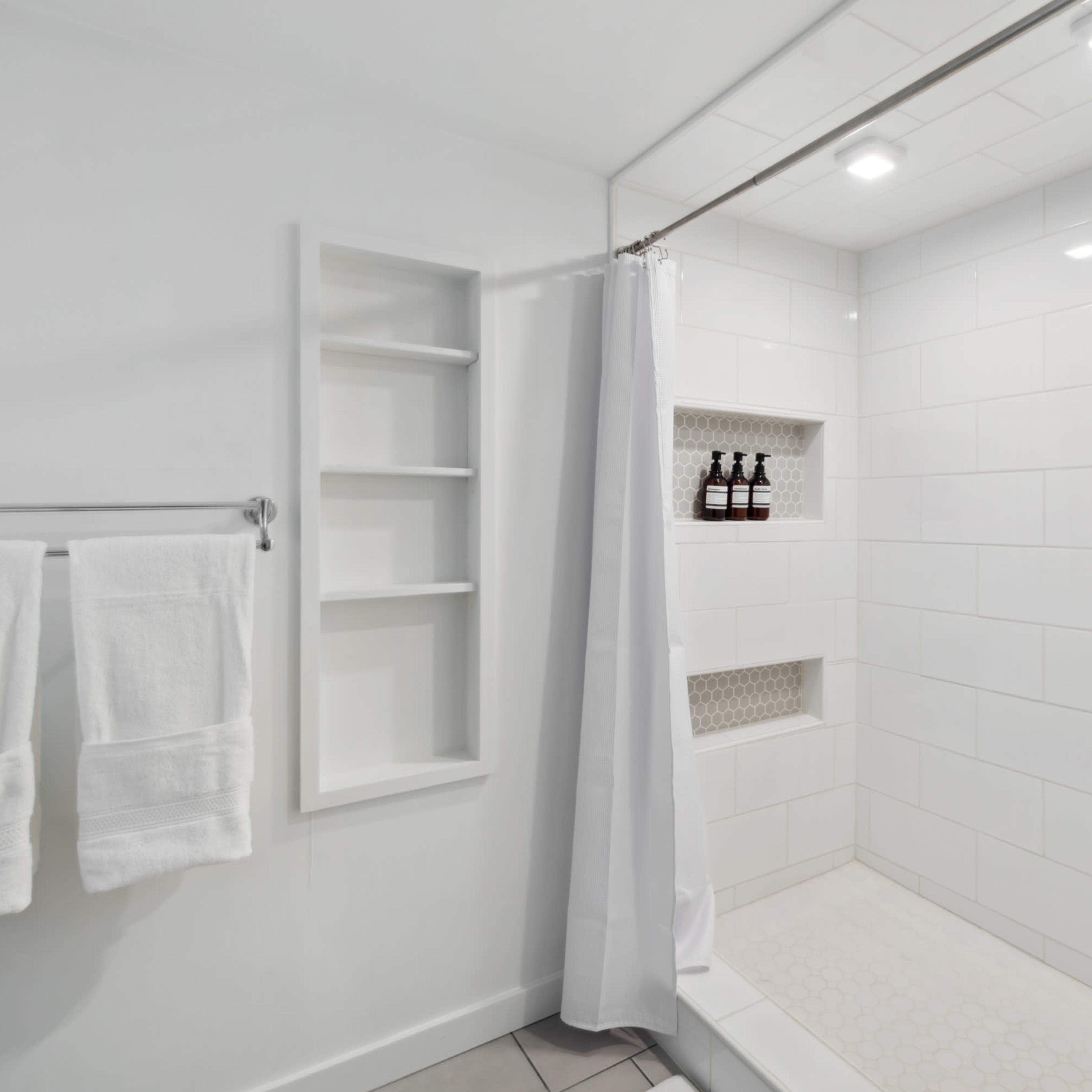 A modern shower with a white tiled wall, a shower curtain, and a recessed shelf displaying several bottles.