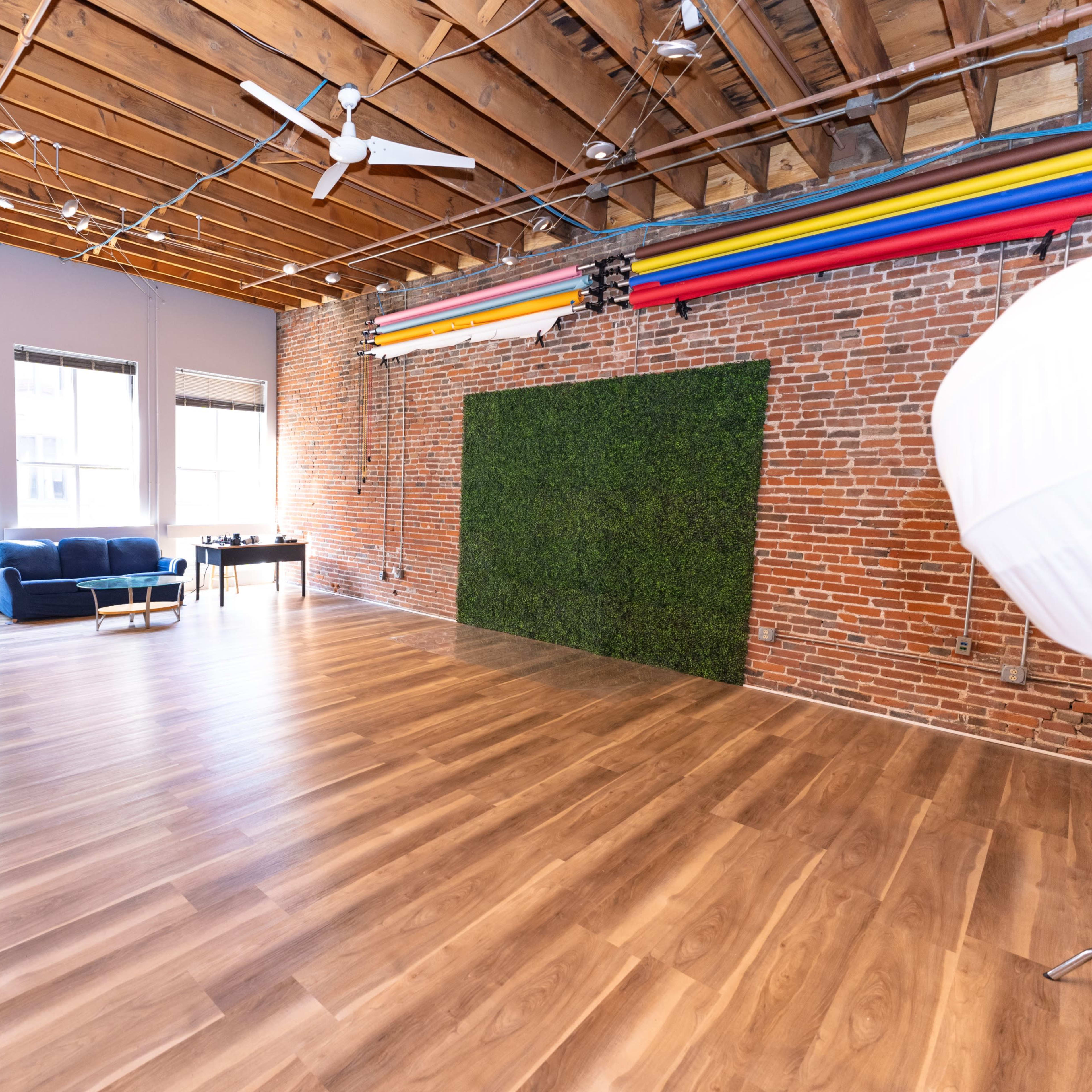 A spacious studio with wooden flooring, exposed brick walls, and a green backdrop, along with photography equipment.
