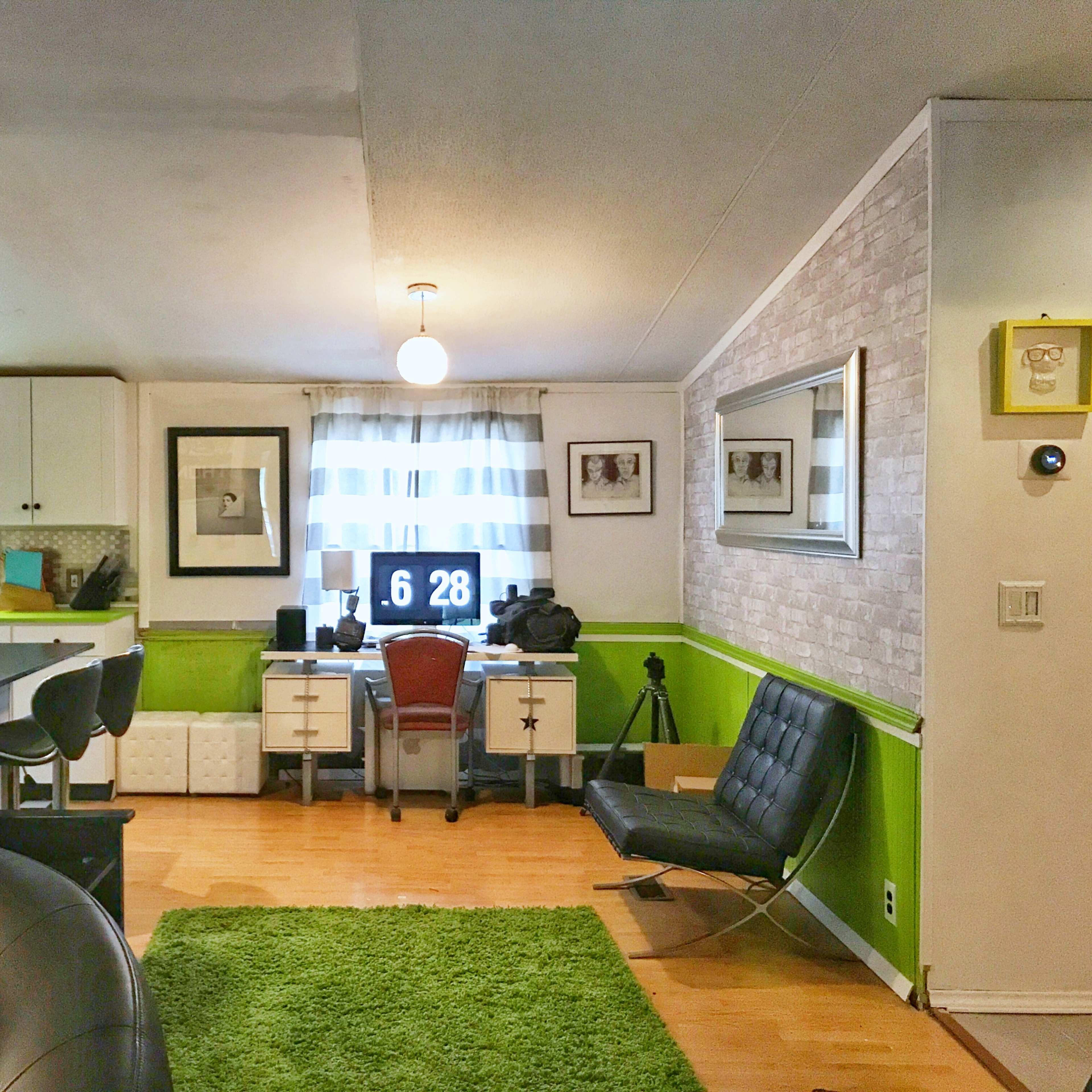 A modern living area with a workspace featuring a desk, computer, and various decorations, along with a green accent wall and a seating area with a black chair.