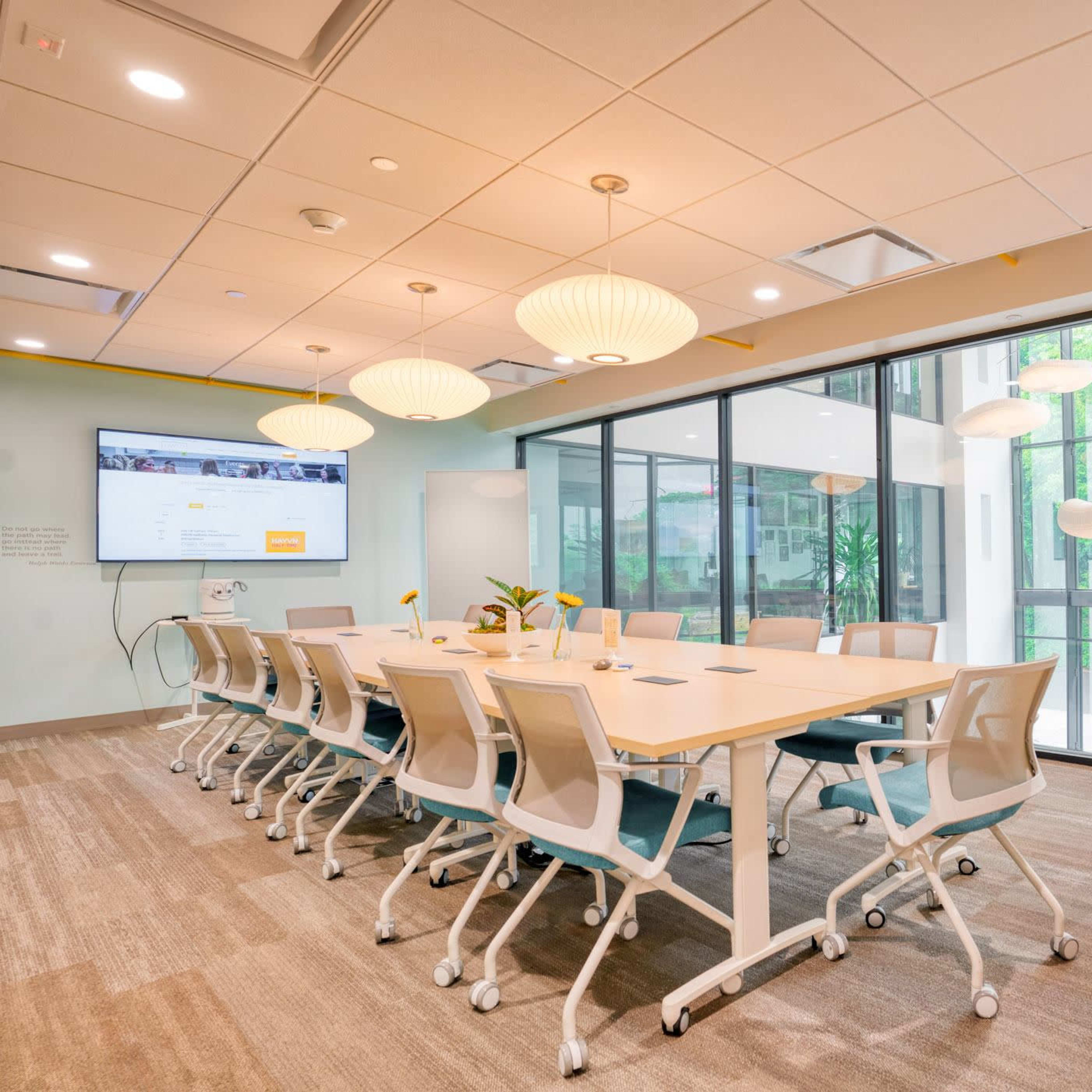 A modern conference room features a large white table surrounded by ergonomic chairs, with floor-to-ceiling windows overlooking greenery and a presentation screen on the wall.