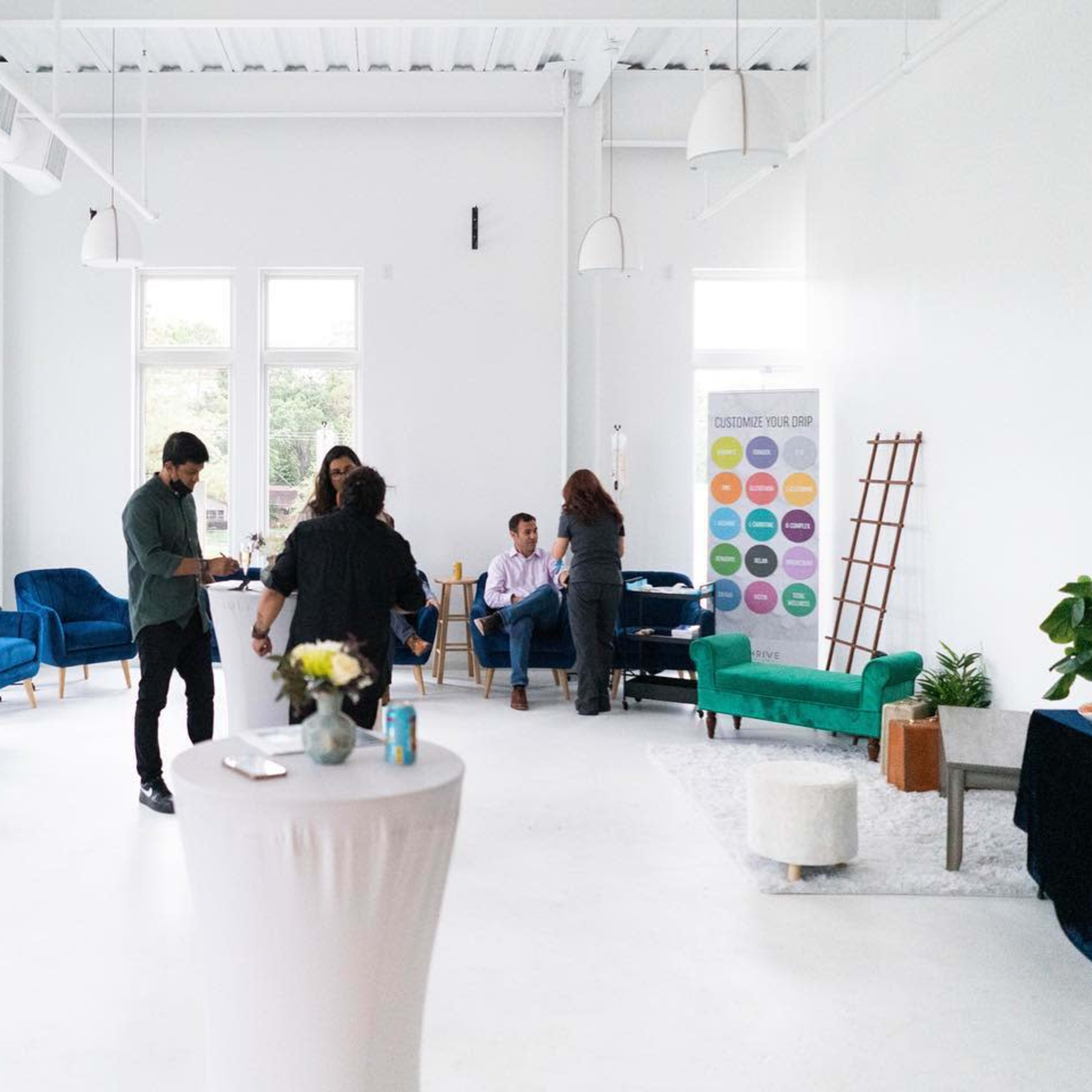 A bright, modern event space features people mingling around tables and plush blue chairs, with large windows allowing natural light to fill the room.