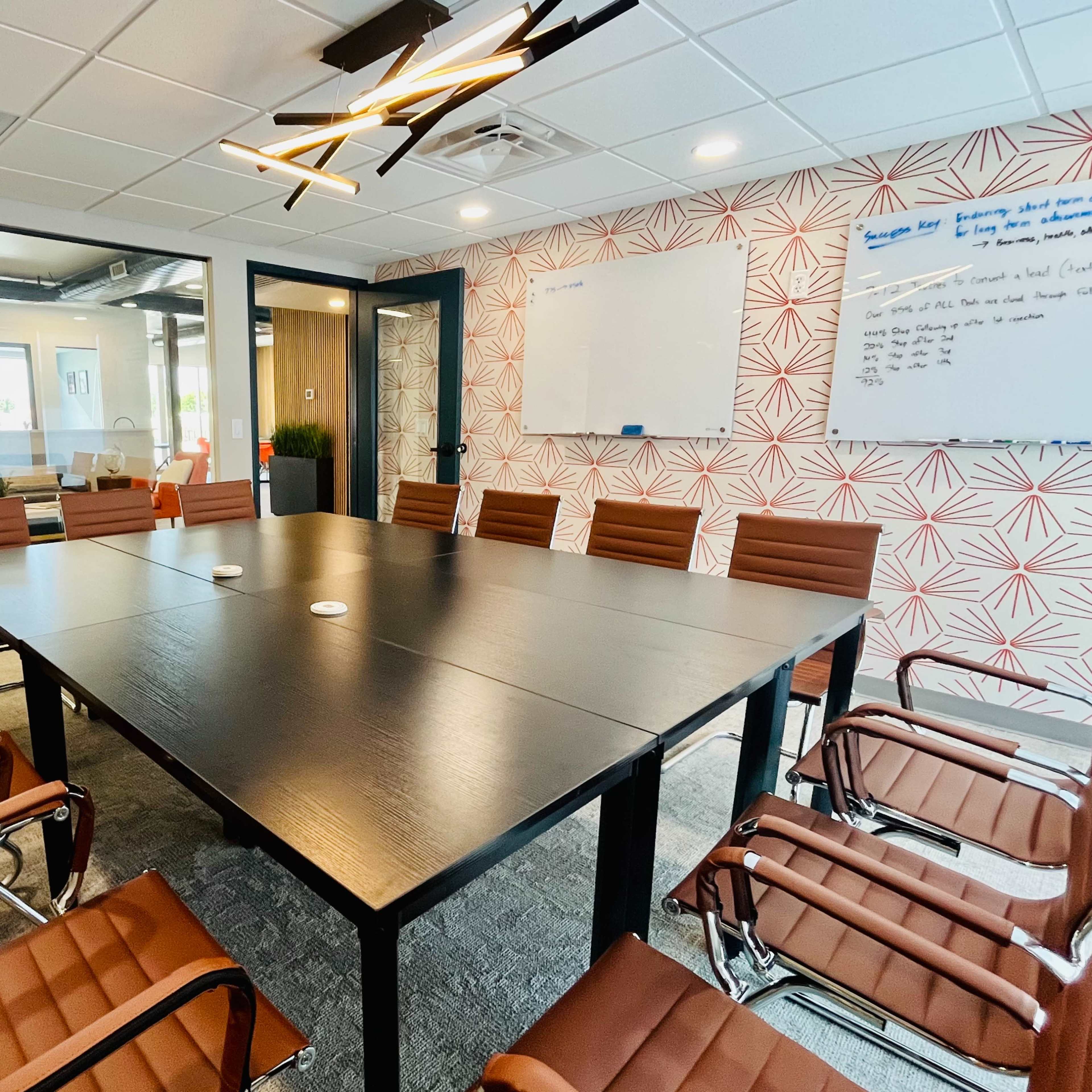 A modern conference room with a large black table surrounded by brown leather chairs, a whiteboard, and patterned wallpaper.