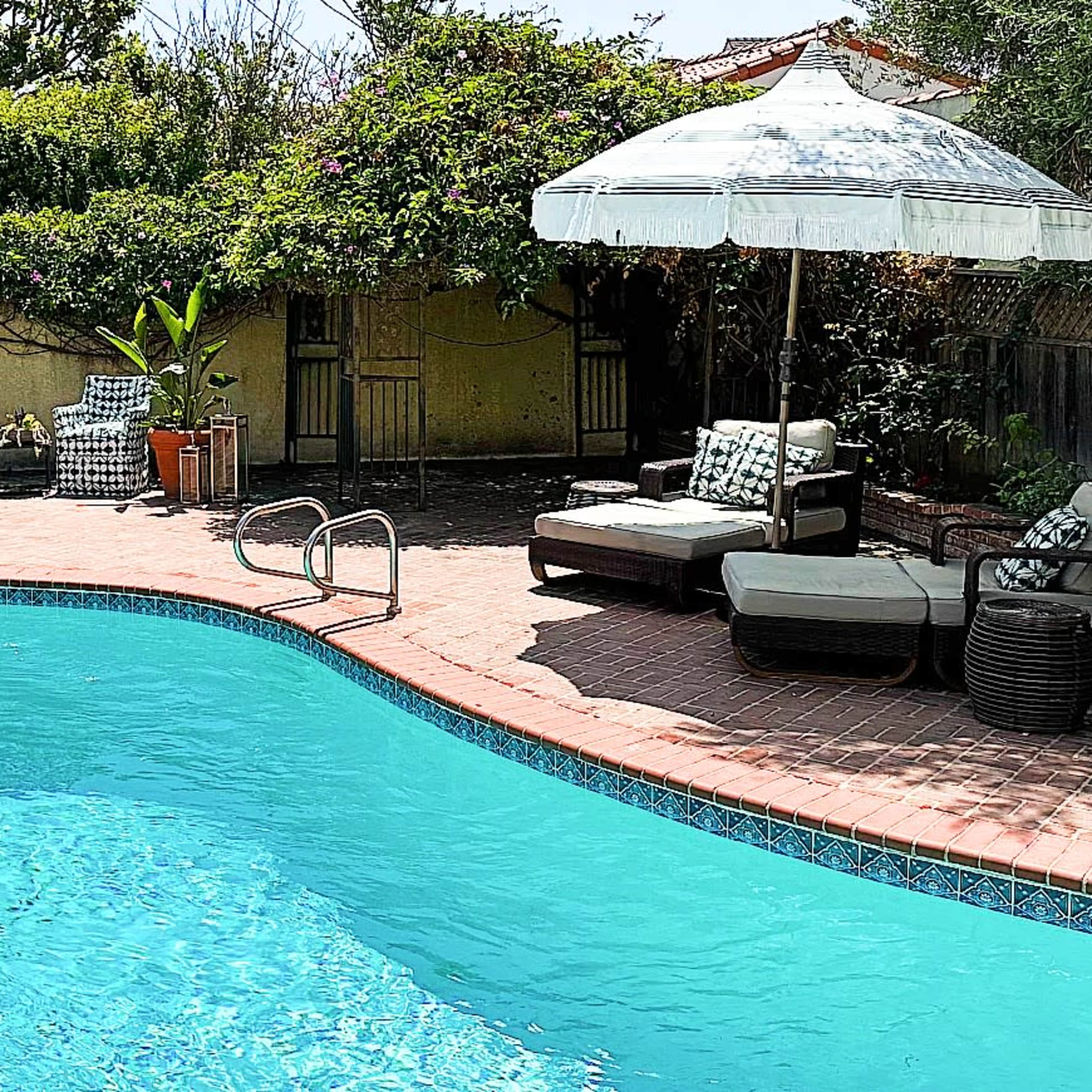A landscaped backyard pool area featuring a blue swimming pool, two lounge chairs, and shaded umbrellas surrounded by greenery.