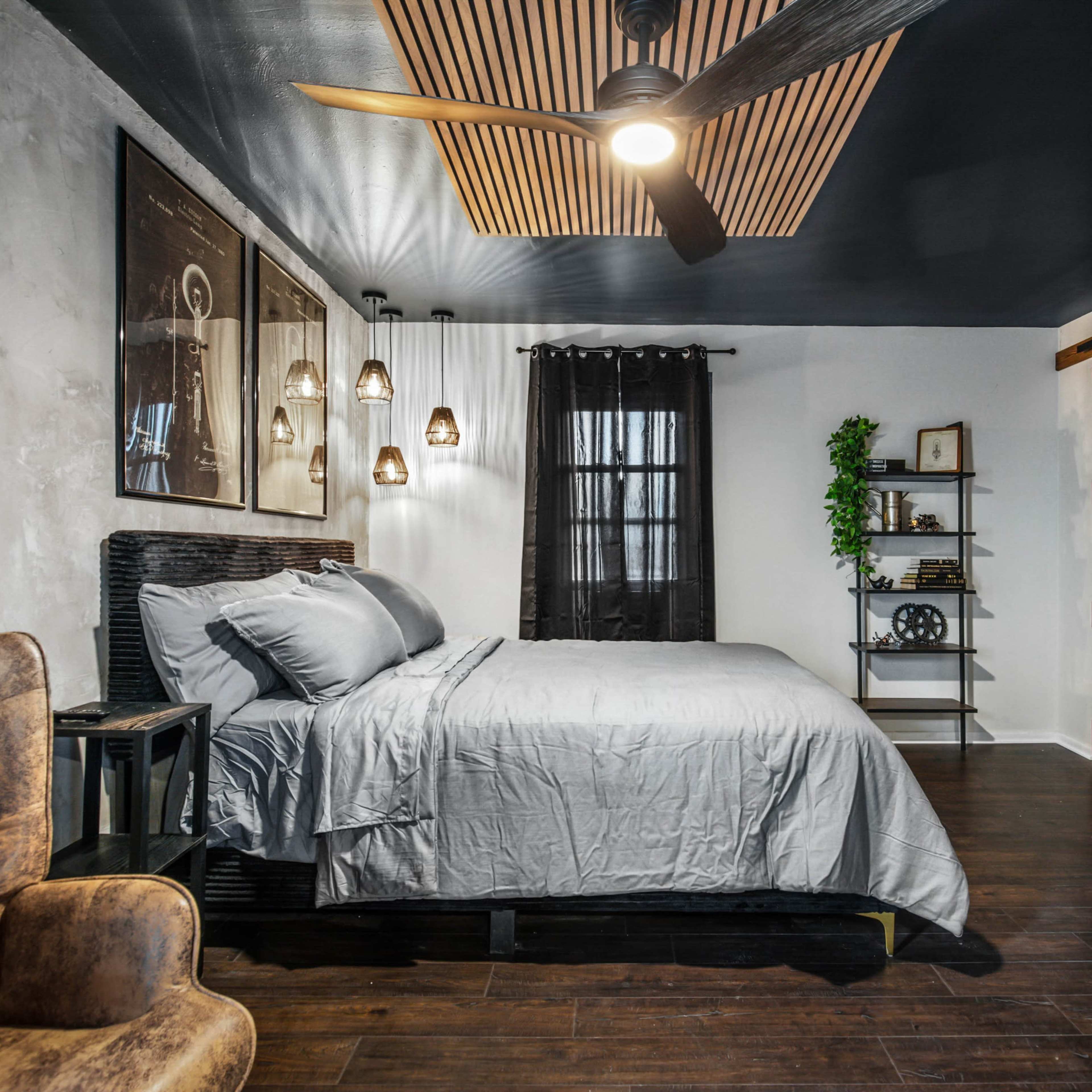 A modern bedroom with a gray bedspread, wooden accent walls, and a ceiling fan, featuring a cozy seating area and a small bookshelf.
