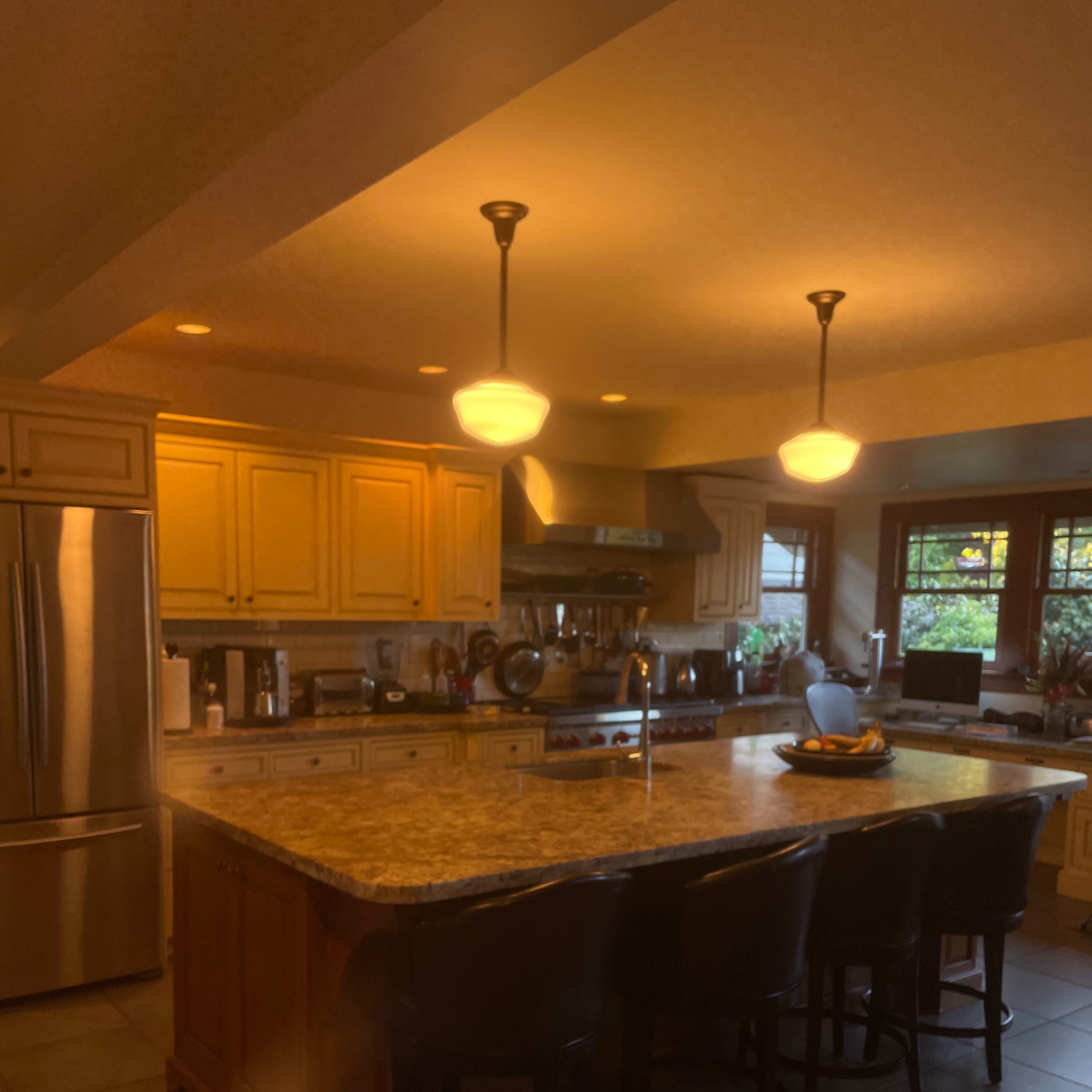 The kitchen features a large island with barstools, wooden cabinetry, and a stainless steel refrigerator, illuminated by pendant lights.