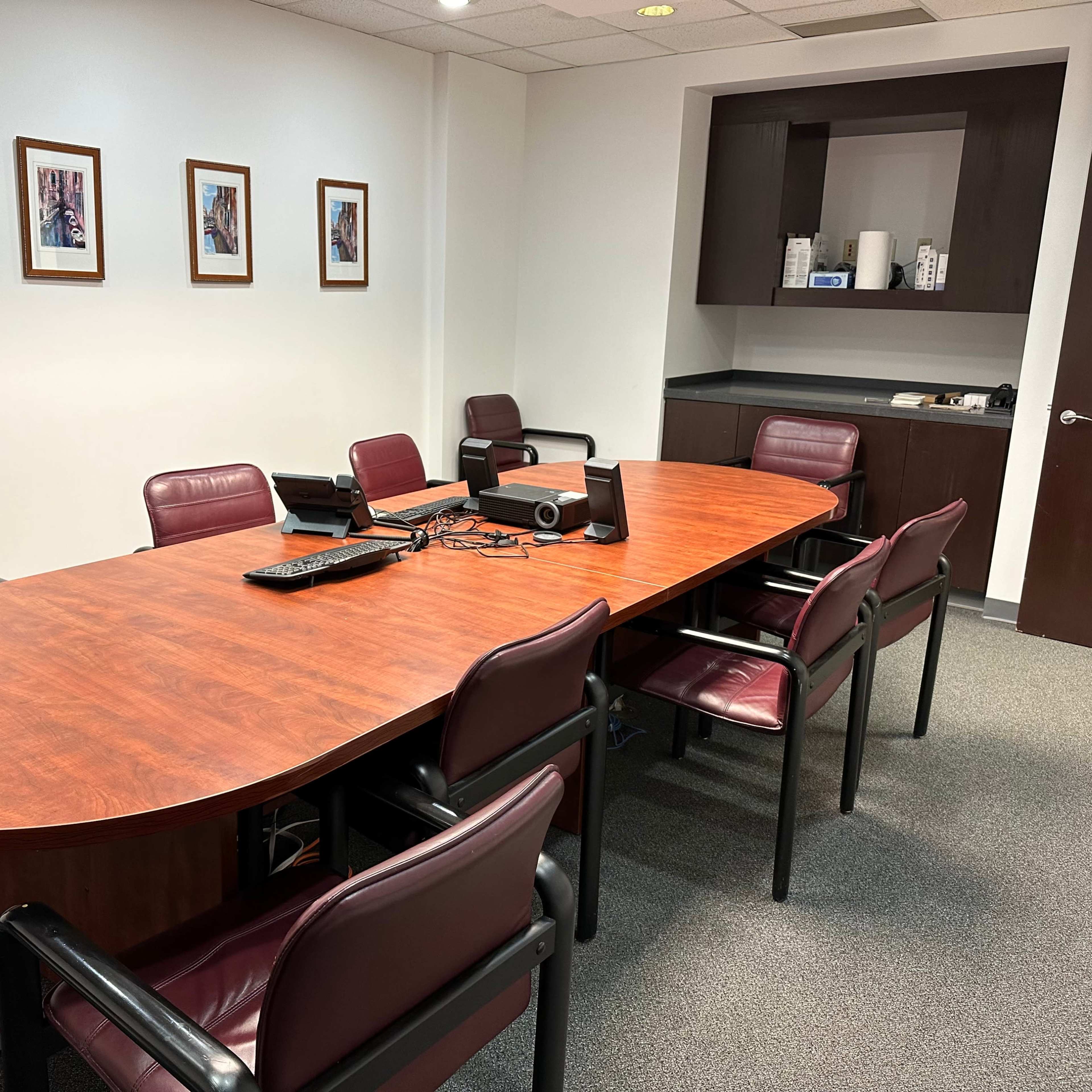 A conference room with an oval wooden table surrounded by six maroon chairs, framed pictures on the walls, and a black cabinet in the corner.