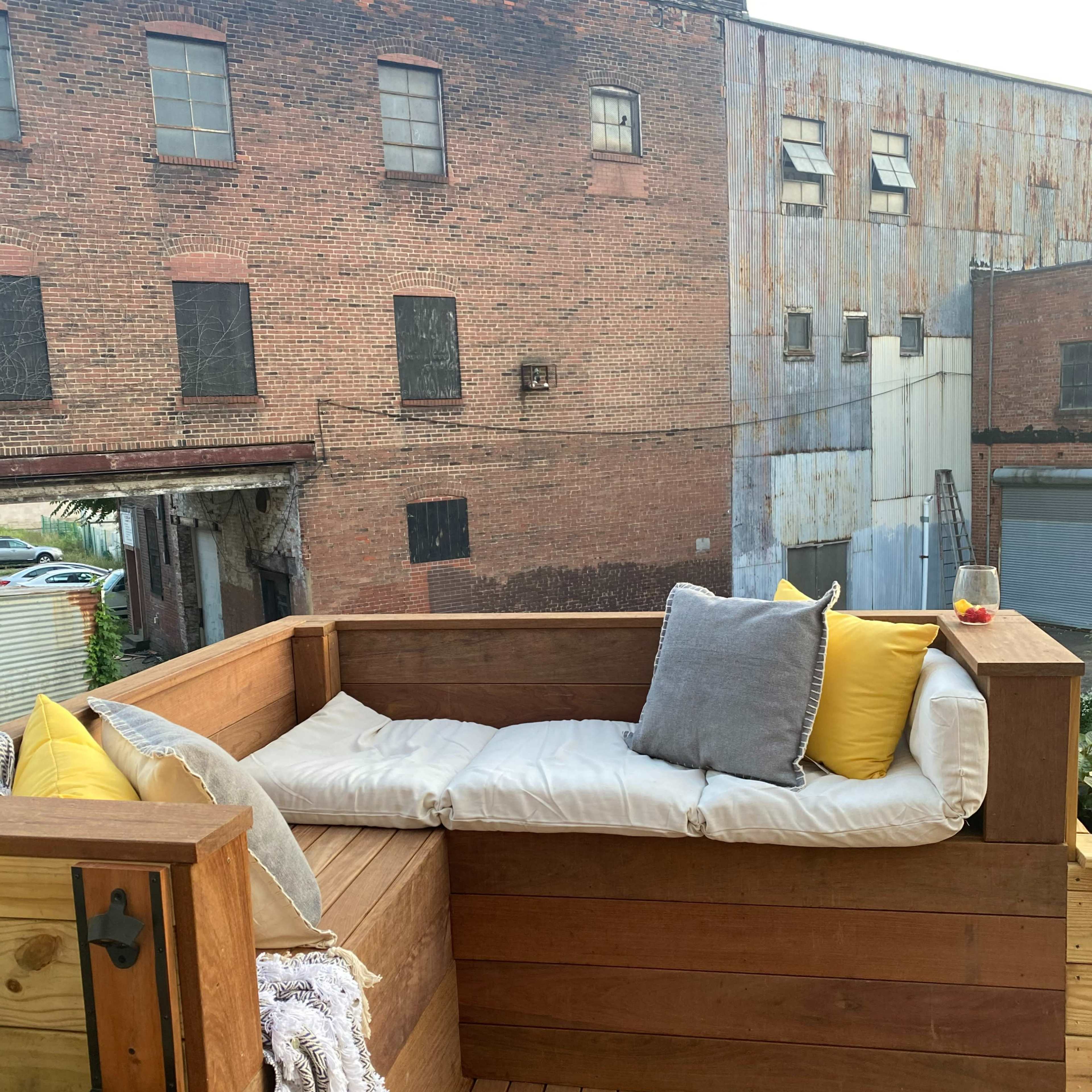 A wooden seating area with cushions and pillows, positioned on a patio overlooking an old brick building.