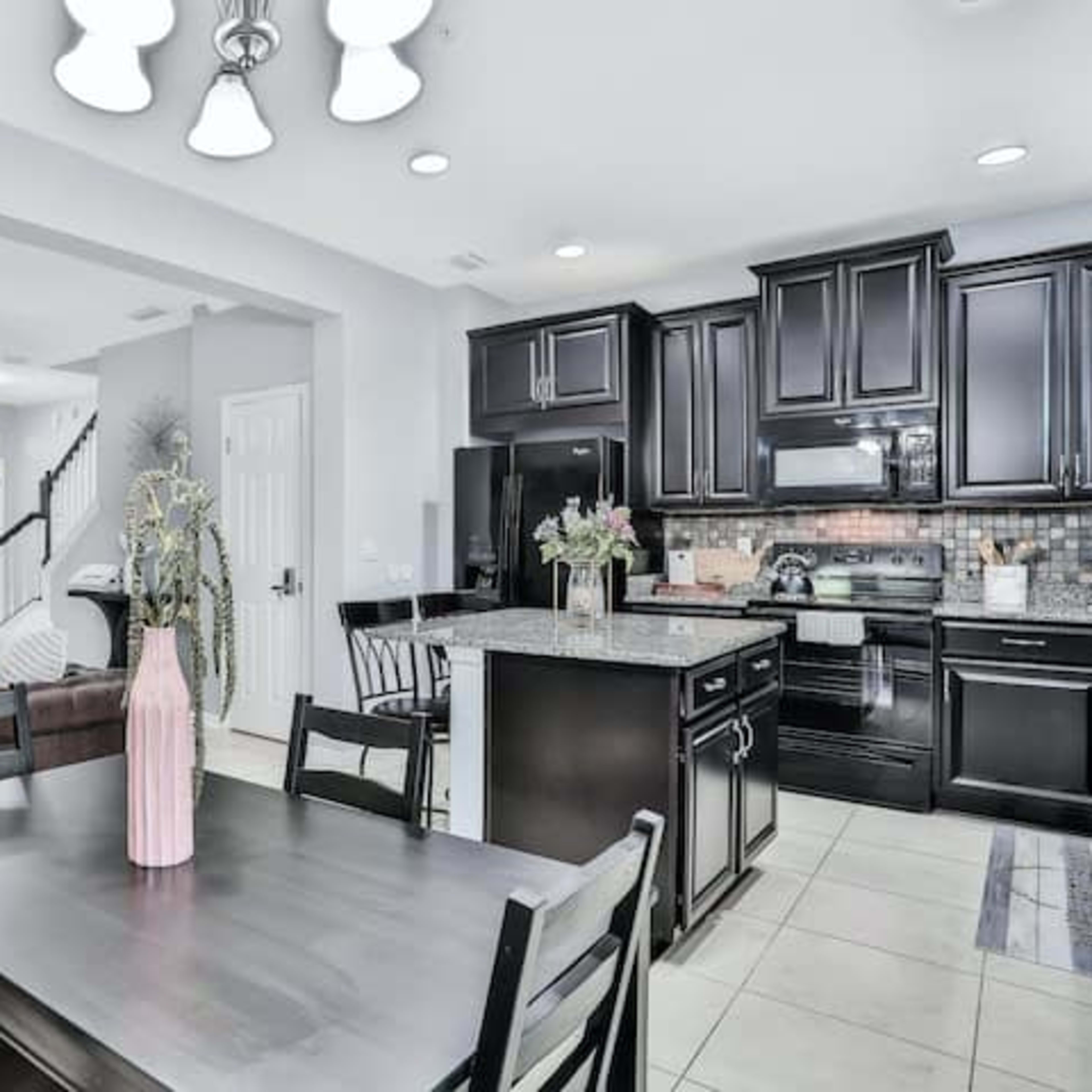 A modern open-concept kitchen and dining area features dark cabinetry, a granite countertop, and a large dining table with a vase of flowers.
