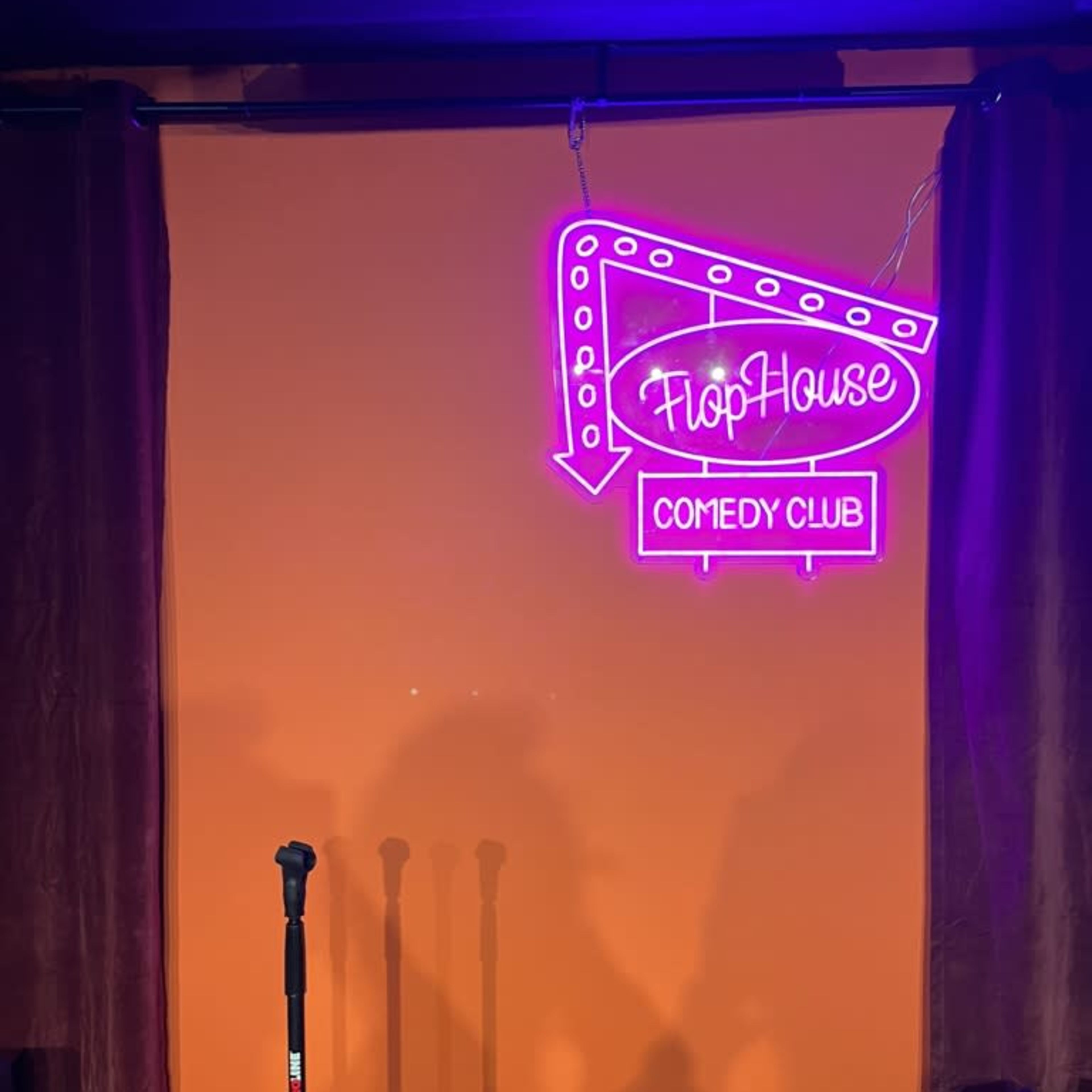 A neon sign for "FlopHouse Comedy Club" illuminated against an orange wall, with a microphone stand in front of it.