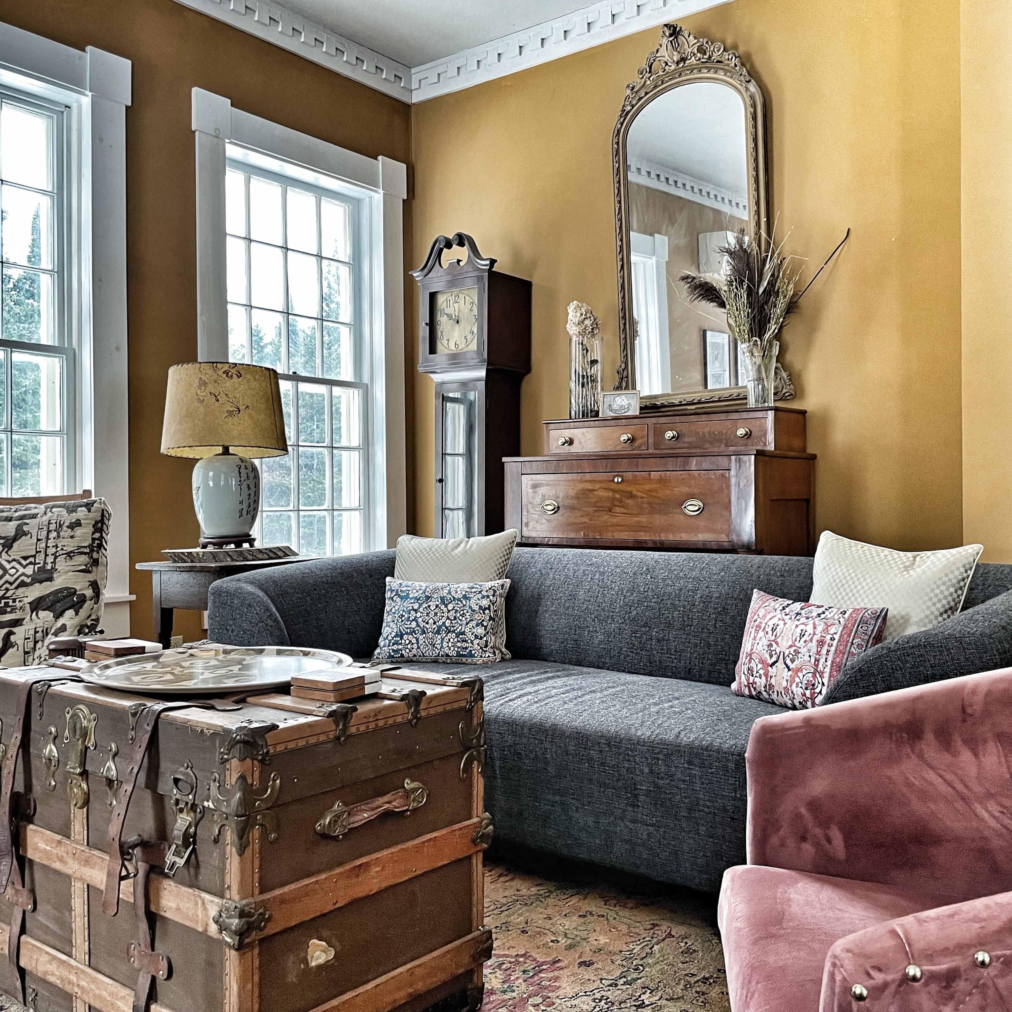 The living room features a gray sofa with decorative pillows, a vintage trunk table, a wooden dresser with a mirror, and large windows allowing natural light to fill the space.