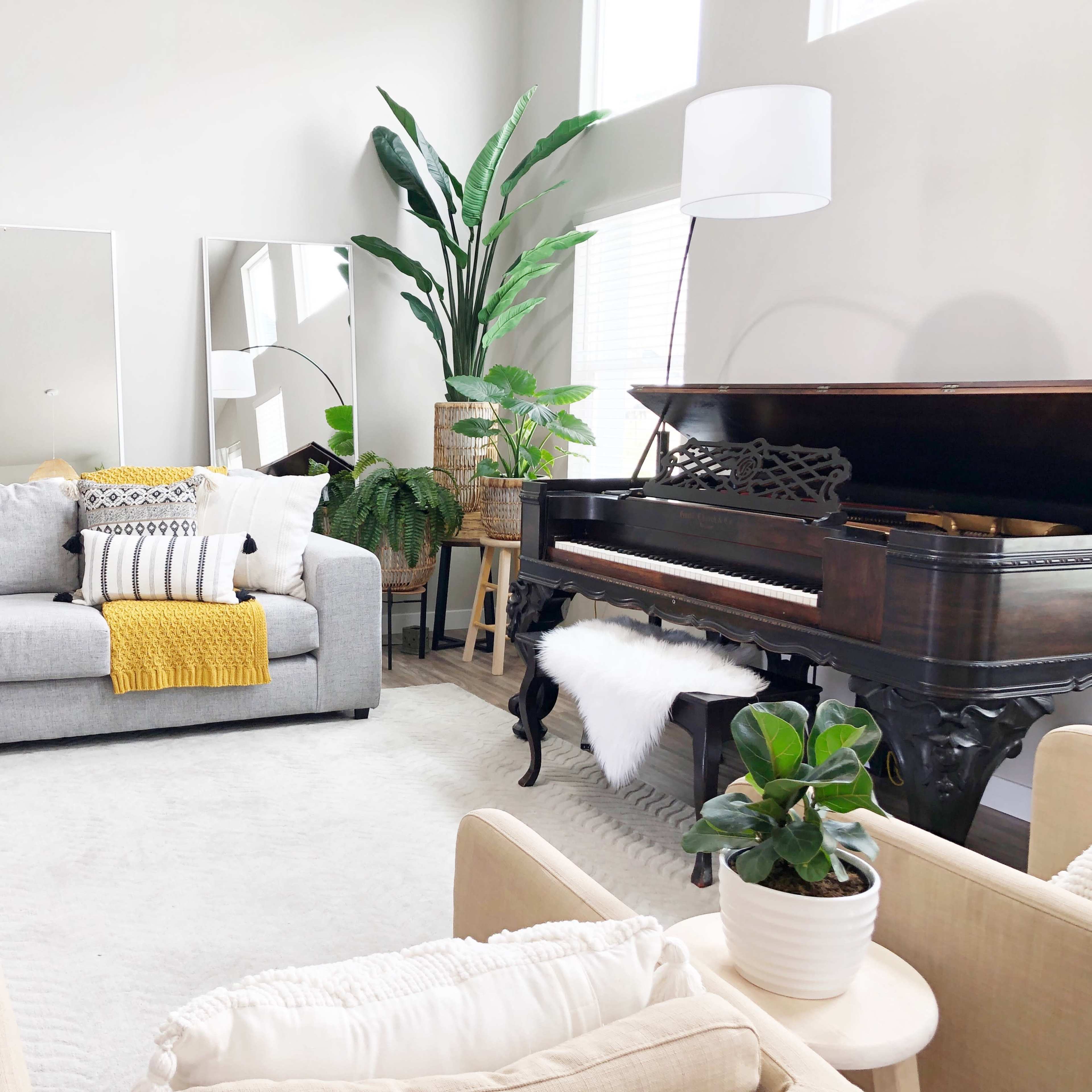 A gray sectional sofa with yellow and white pillows is positioned beside a piano and surrounded by plants in a bright living room.