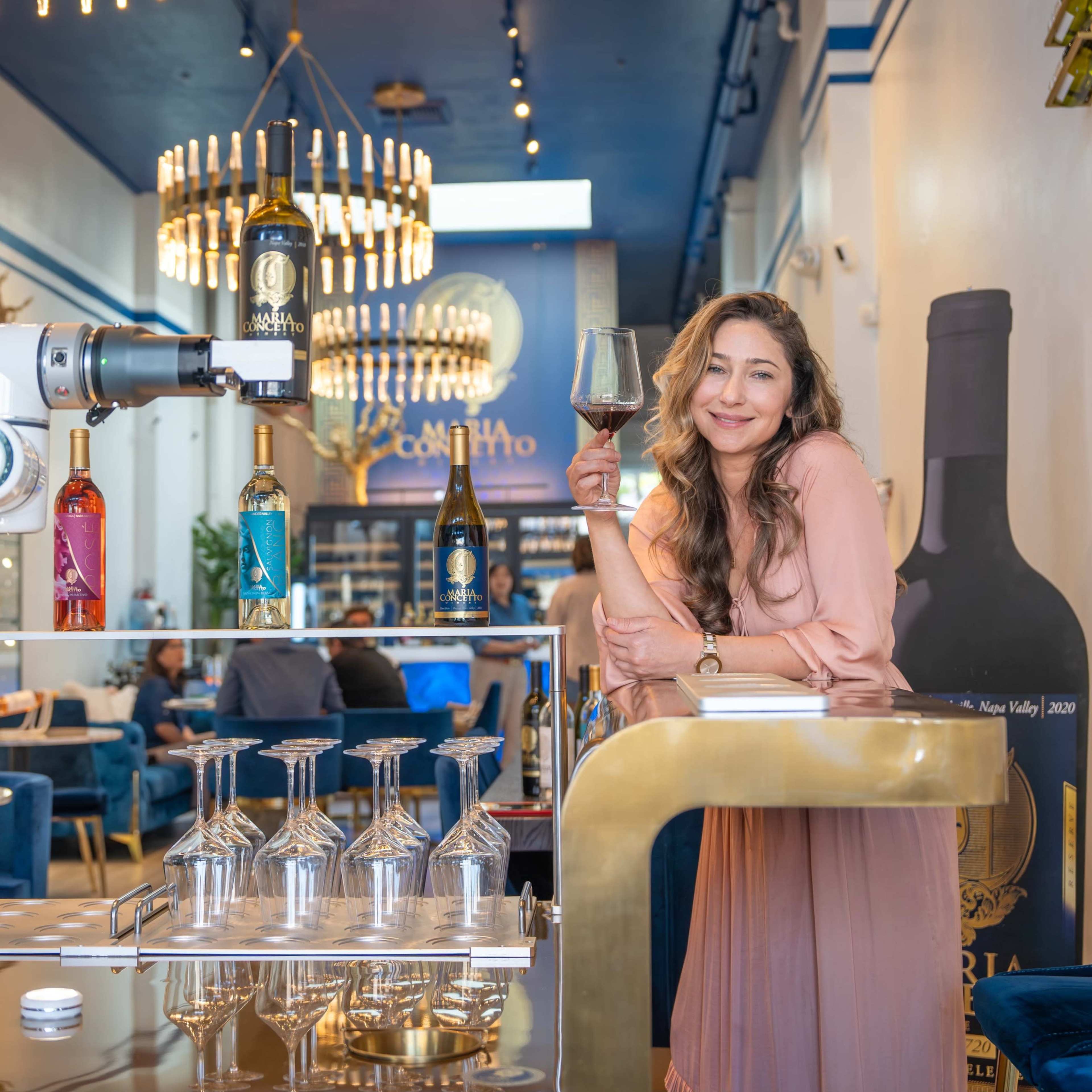 A robotic arm is serving drinks while a woman stands next to a table in a modern bar filled with stylish furniture and decorative elements.