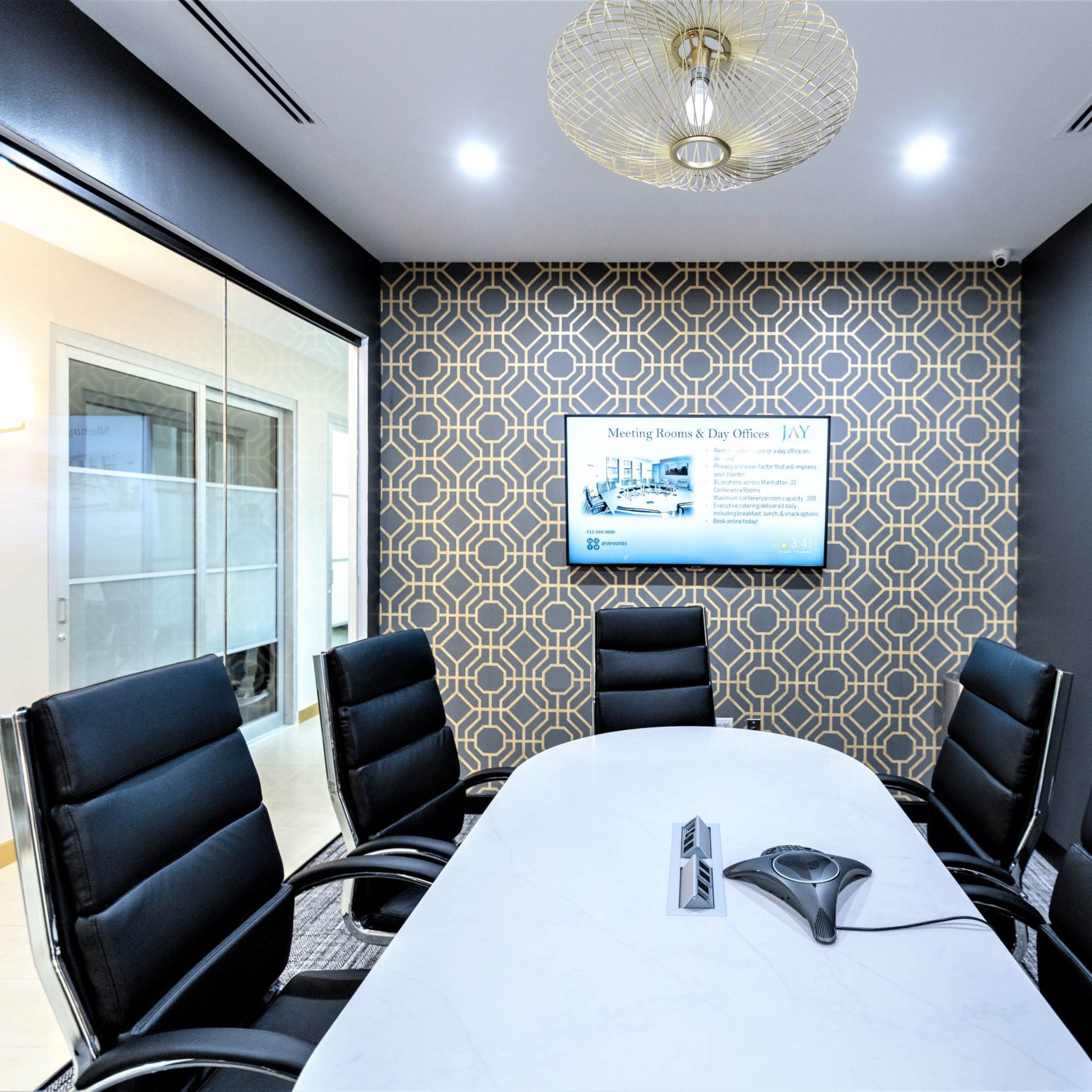 A modern conference room with a large white table, black leather chairs, and a patterned wall featuring a television screen displaying information.