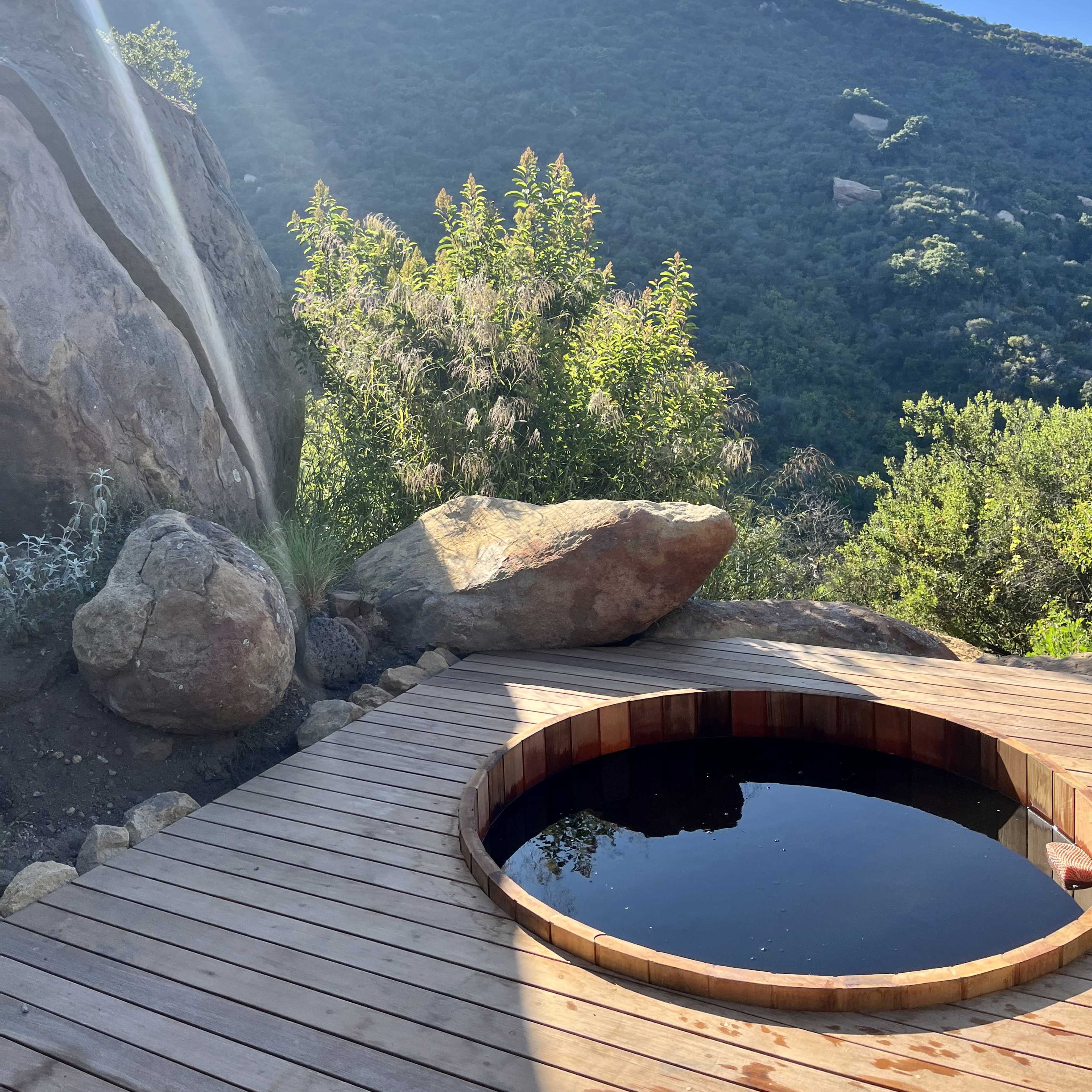 A round wooden hot tub sits on a wooden deck surrounded by greenery and large rocks, with a hillside in the background.