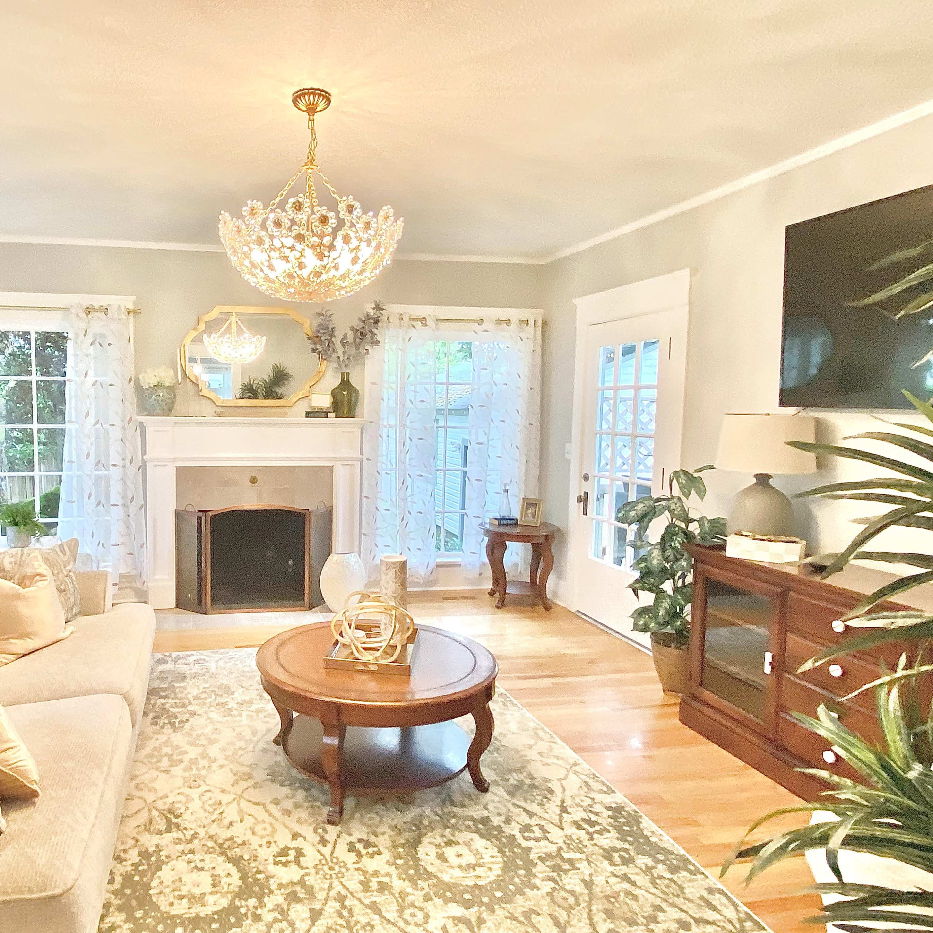 The living room features a beige sofa, a round wooden coffee table, and a large chandelier hanging from the ceiling, with large windows allowing natural light to illuminate the space.