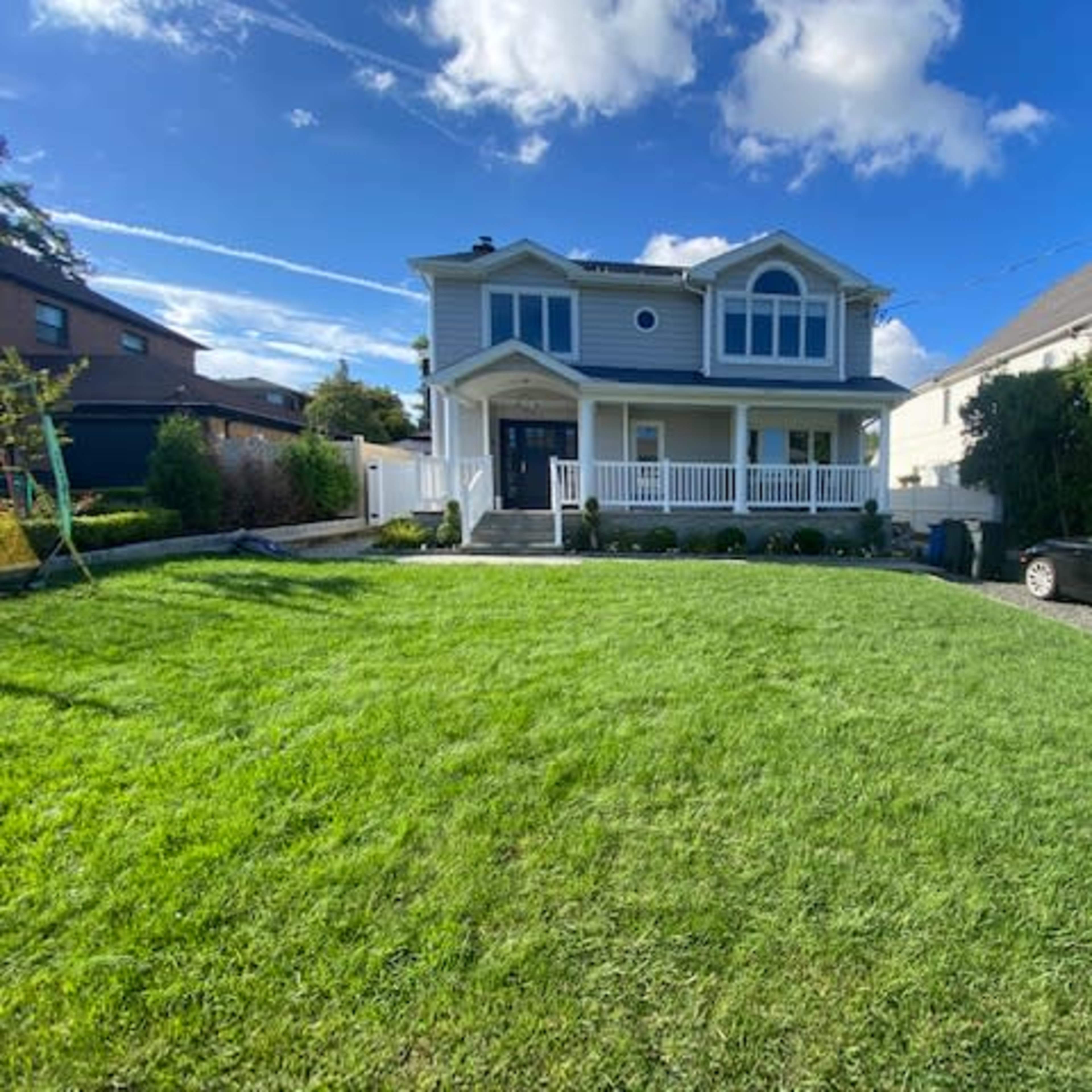 A two-story house with a blue exterior and a front porch is set on a well-maintained lawn, with a black car parked in the driveway.