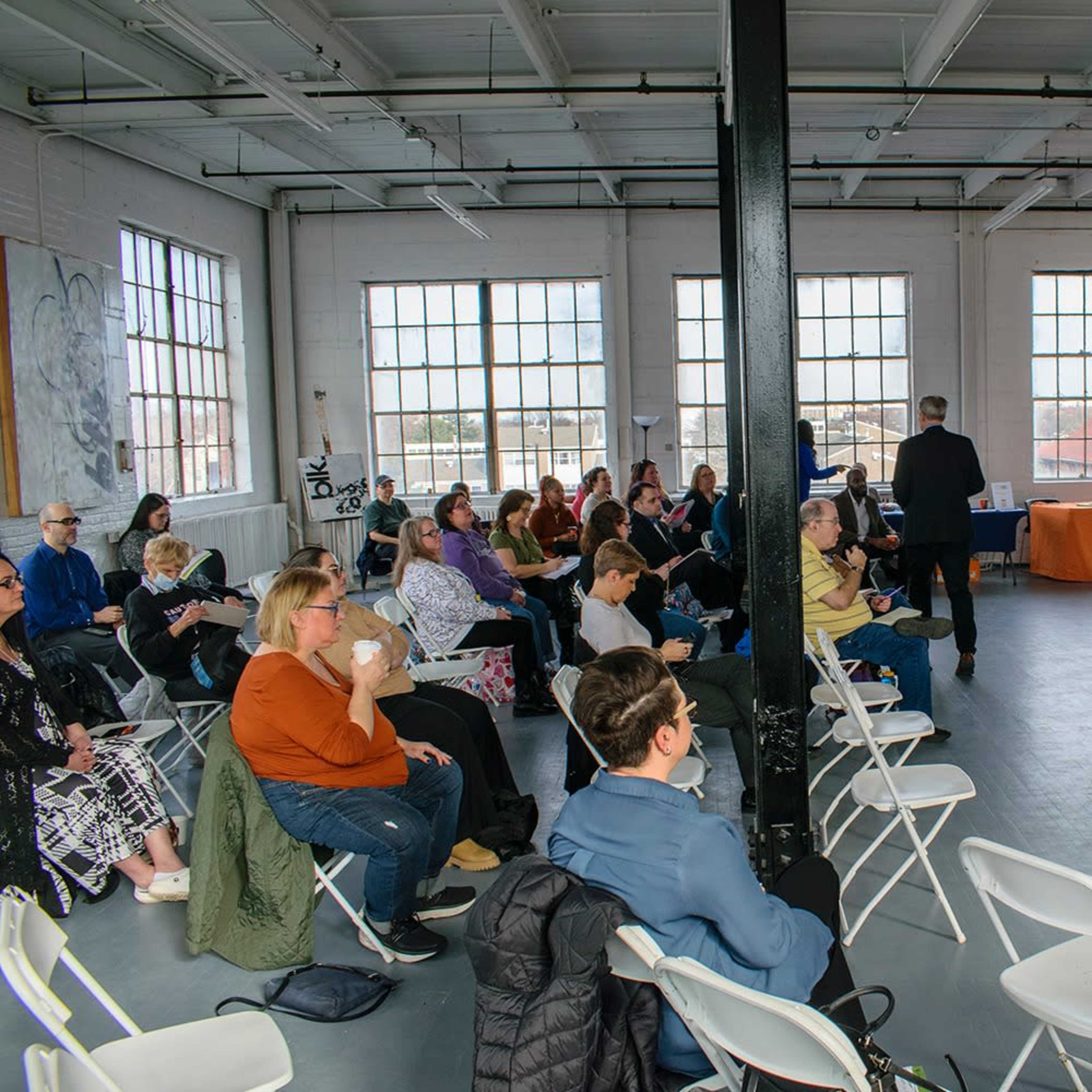 A group of people sits in white chairs arranged in a spacious room with large windows, while a speaker stands at a microphone near an orange table.