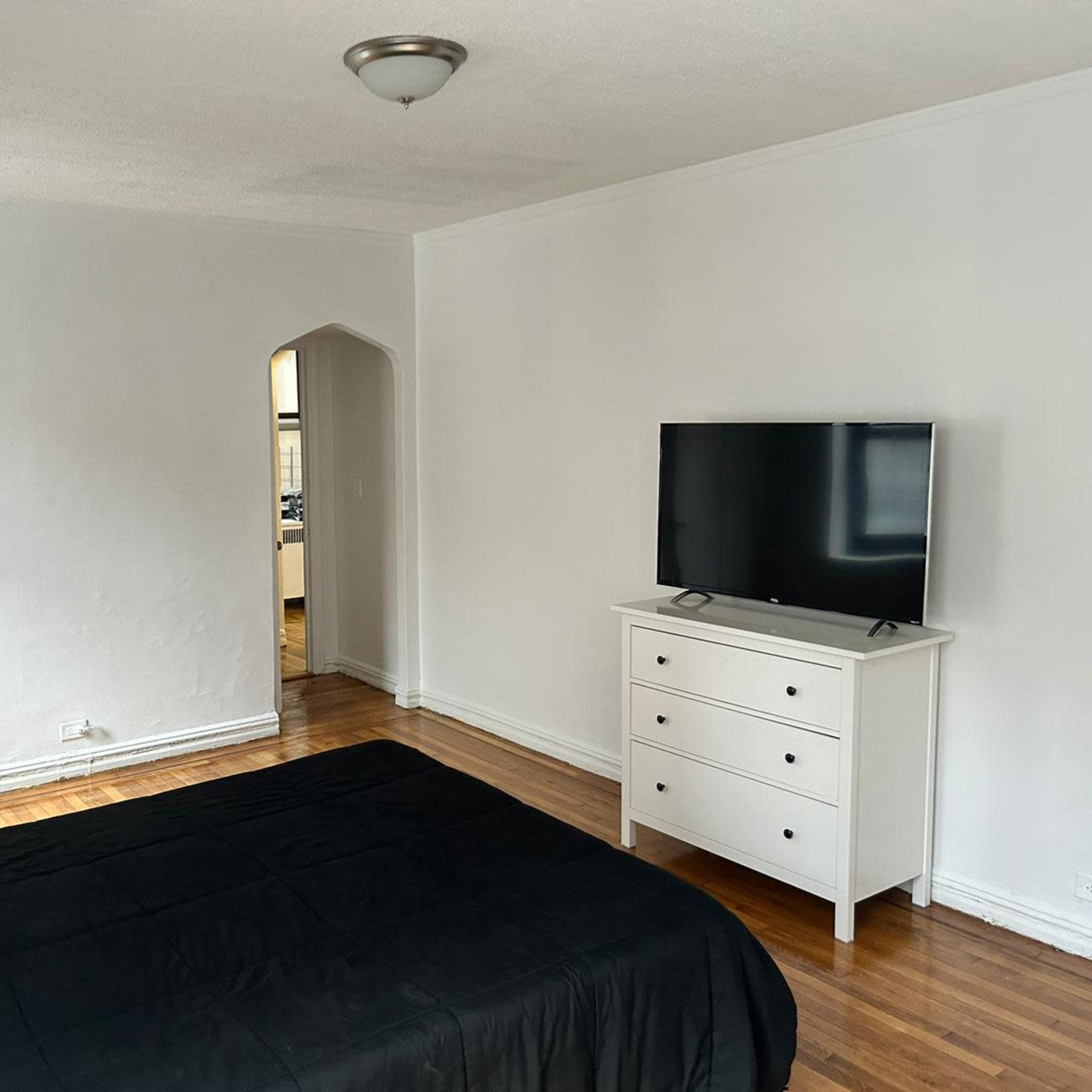 A simple bedroom features a bed with a black bedspread, a white dresser, and a television on a stand against a white wall.