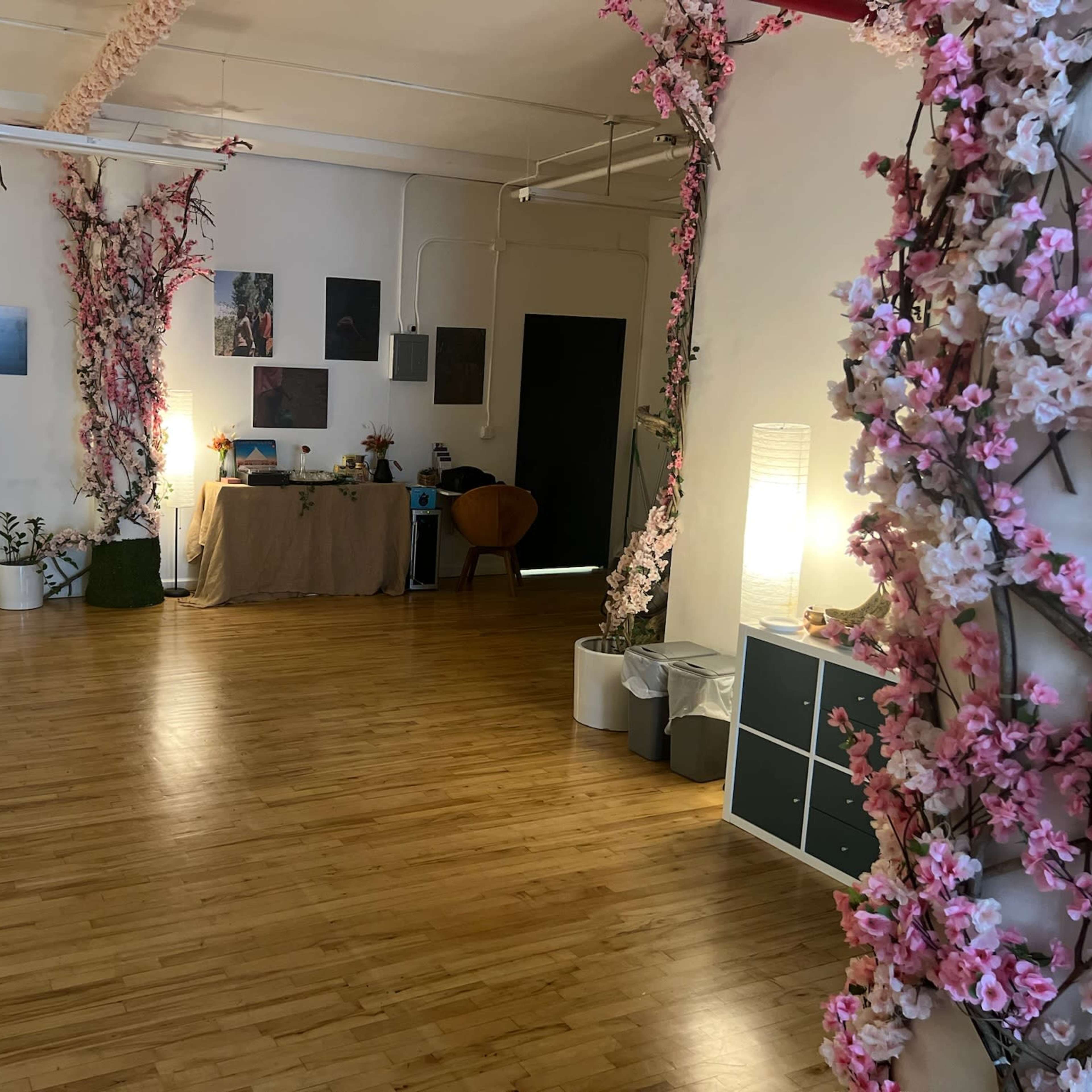 A spacious room with wooden flooring, decorated with pink flower arrangements on either side, and various furniture items including a table, a chair, and storage units.