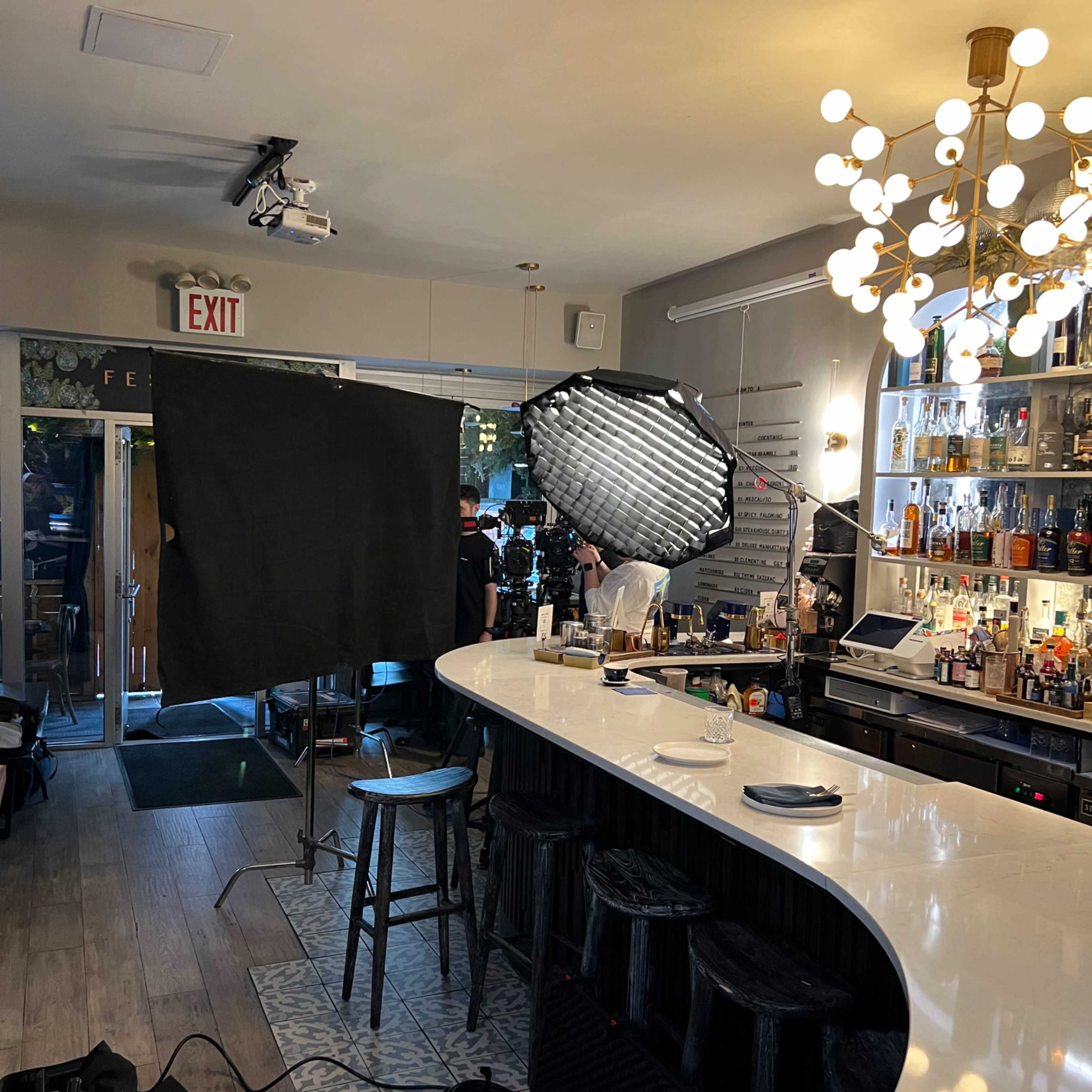 A bar area is set up for a photoshoot, featuring a white countertop, a large umbrella light, and a camera crew in the background.