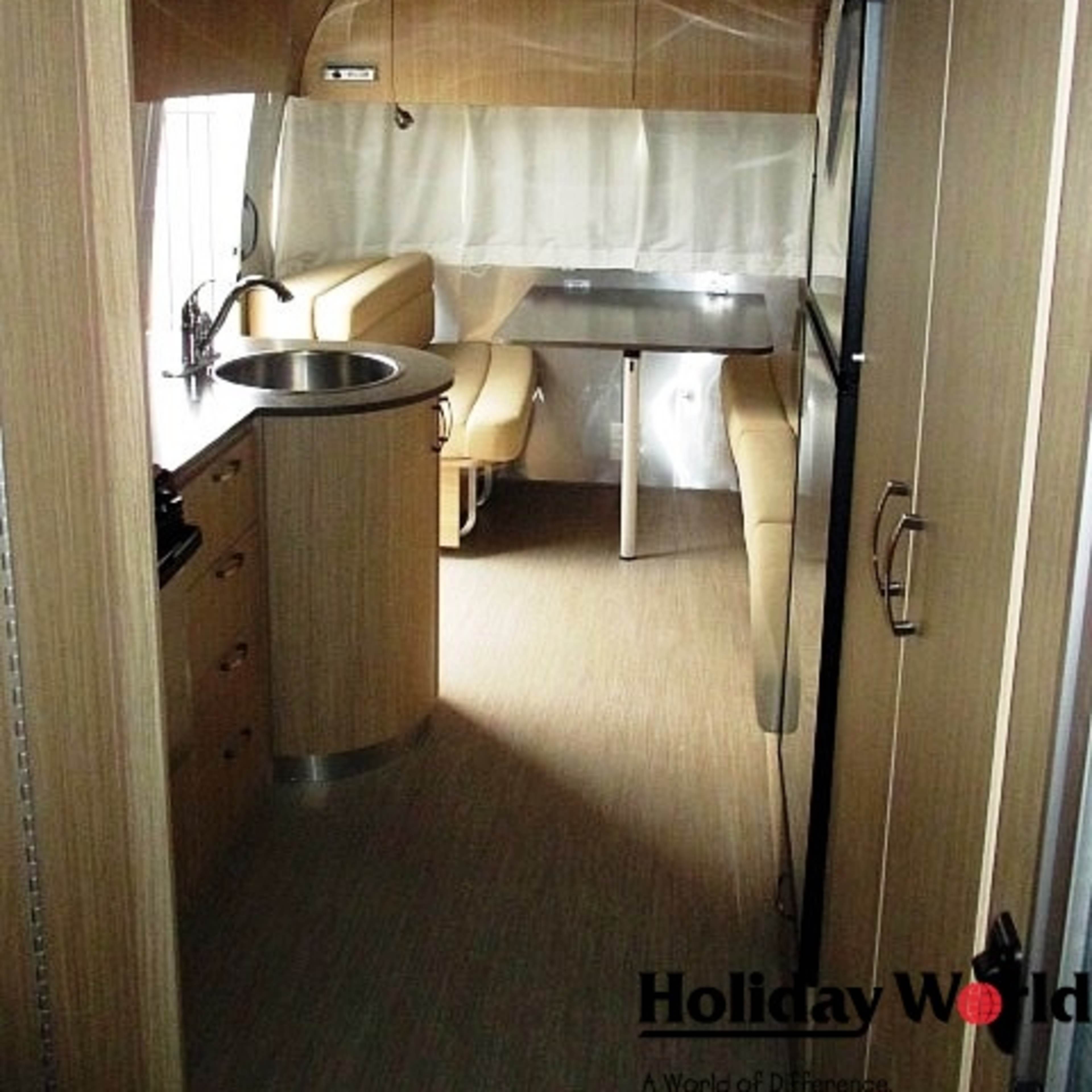 The interior of a streamlined camper or trailer with wood paneling, a kitchenette, dining area, and a central table.