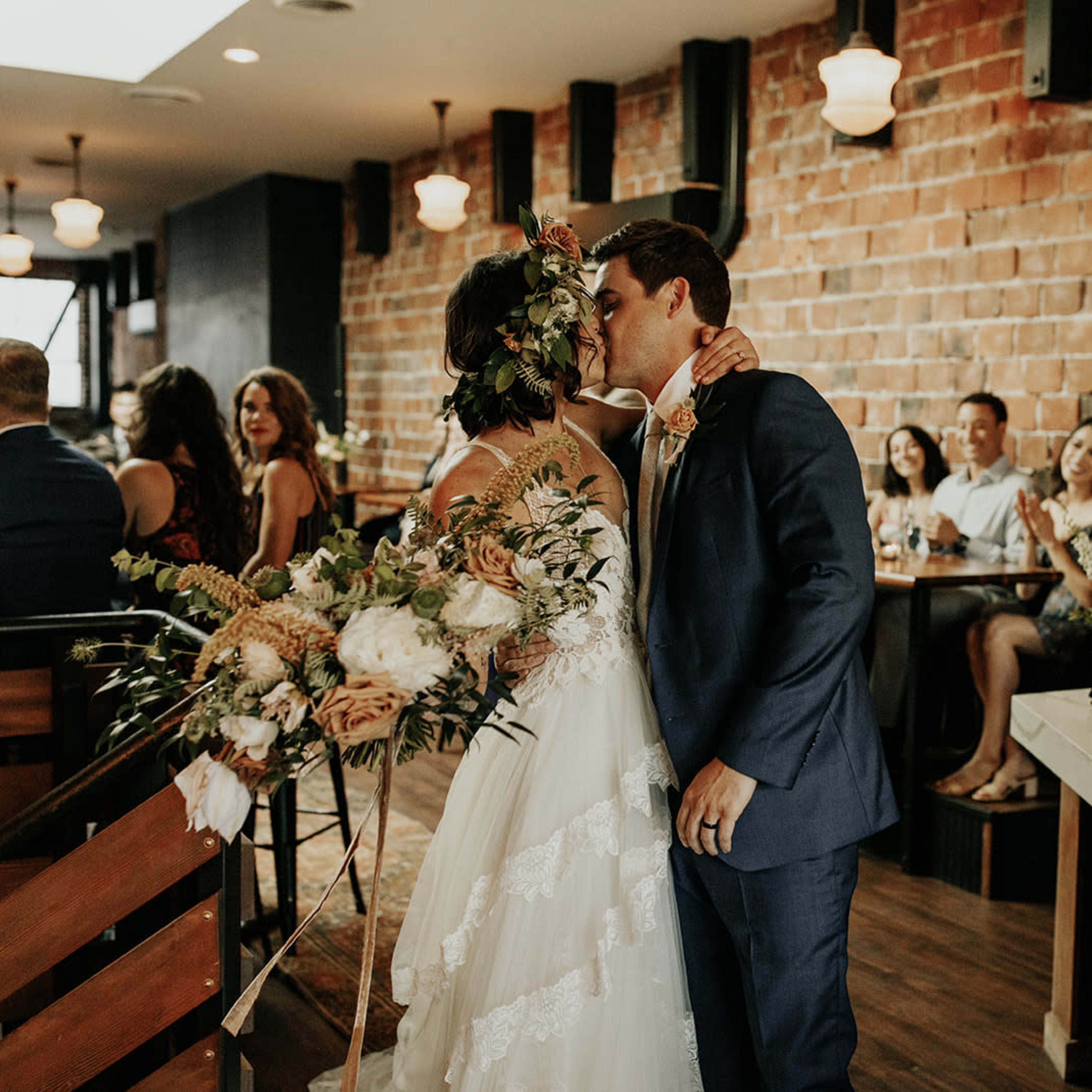 A bride and groom kiss at their wedding reception, surrounded by guests seated at tables adorned with flowers.