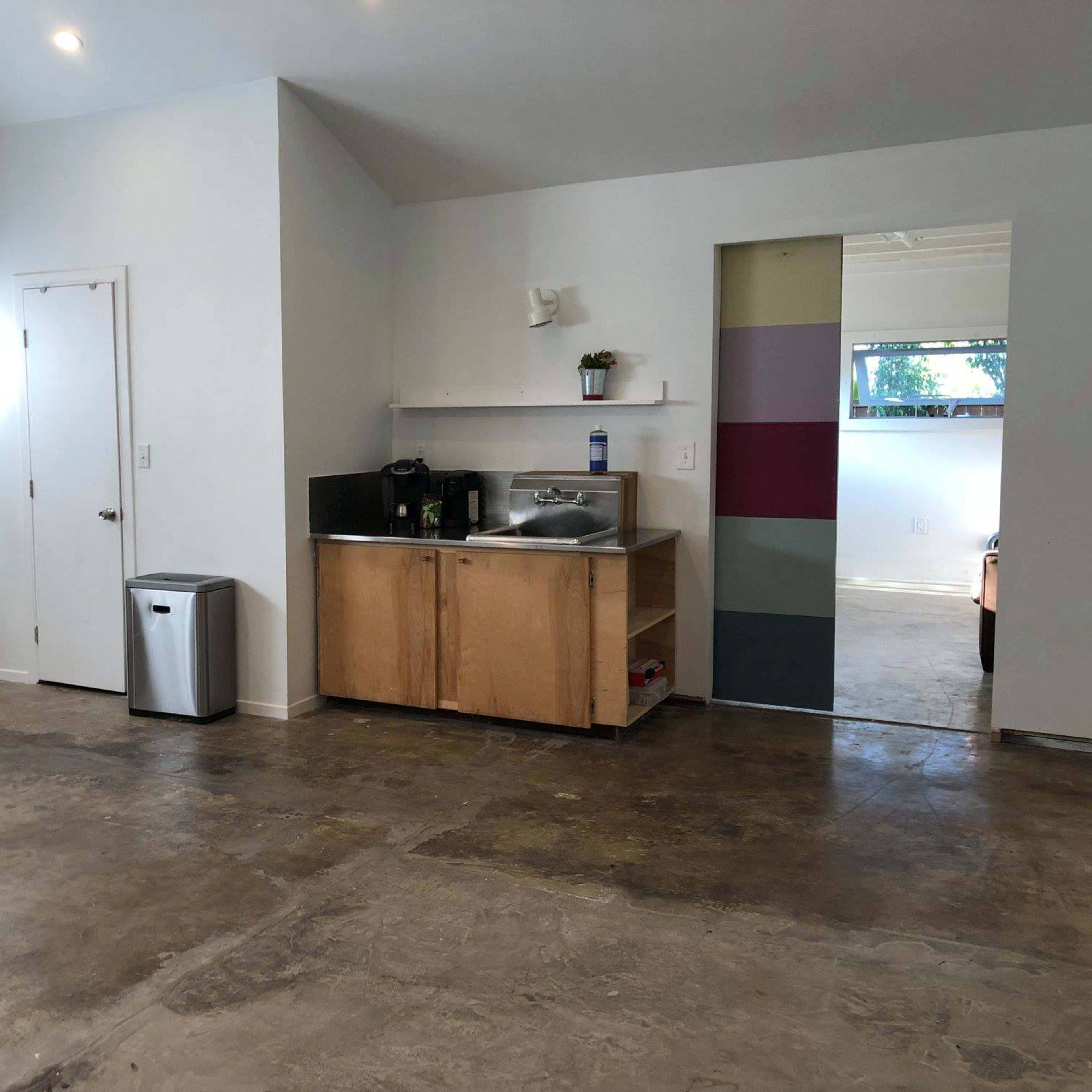 An interior space with a kitchenette, a door leading outside, and a partially visible room through an open doorway.