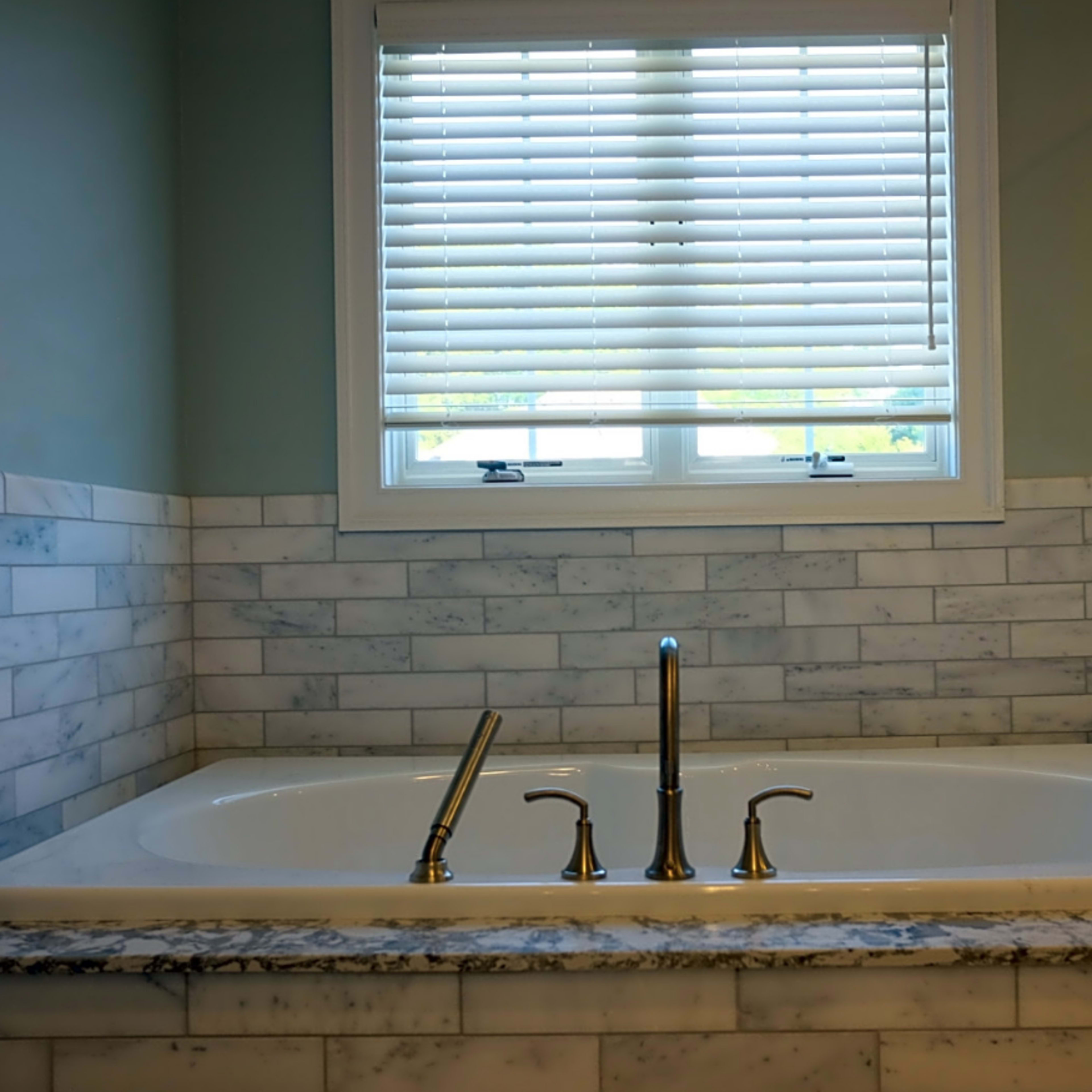 A white bathtub with three faucets, set against a background of marble tiles and a window with white blinds.