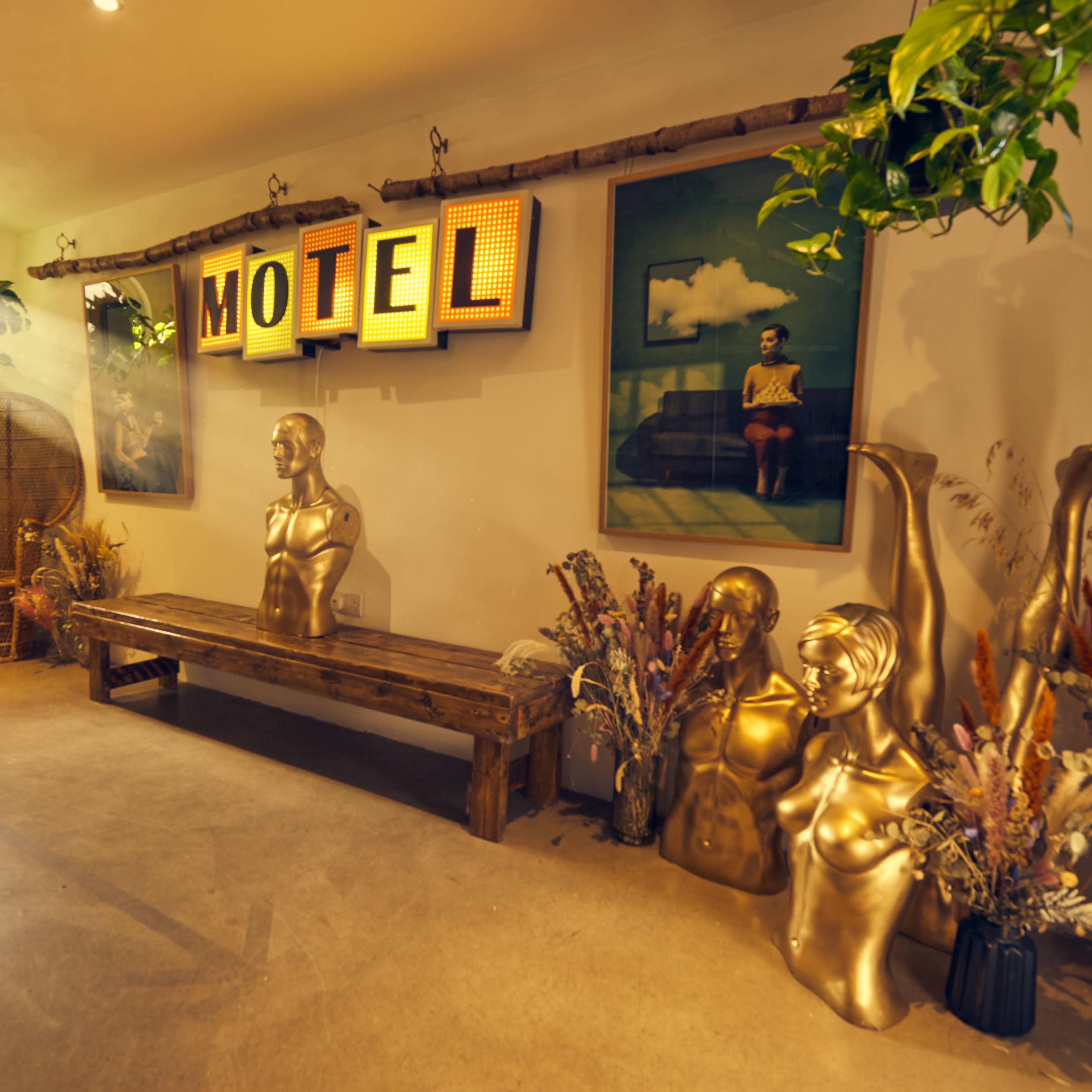 The interior of a stylish motel features two gold mannequins, a wooden bench, and framed art, all surrounded by decorative plants.