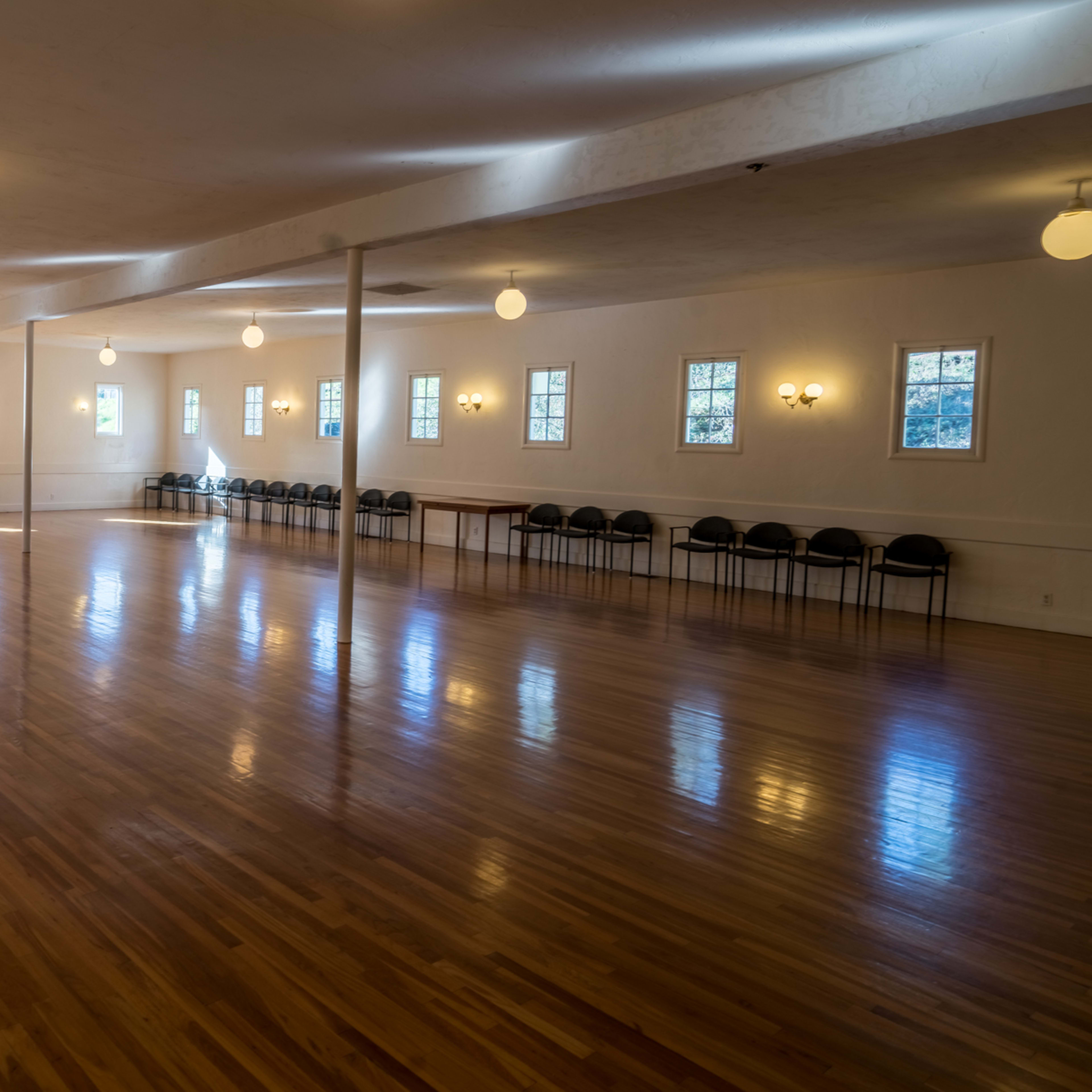 A spacious, brightly lit empty hall with wooden flooring and a row of chairs along one wall.