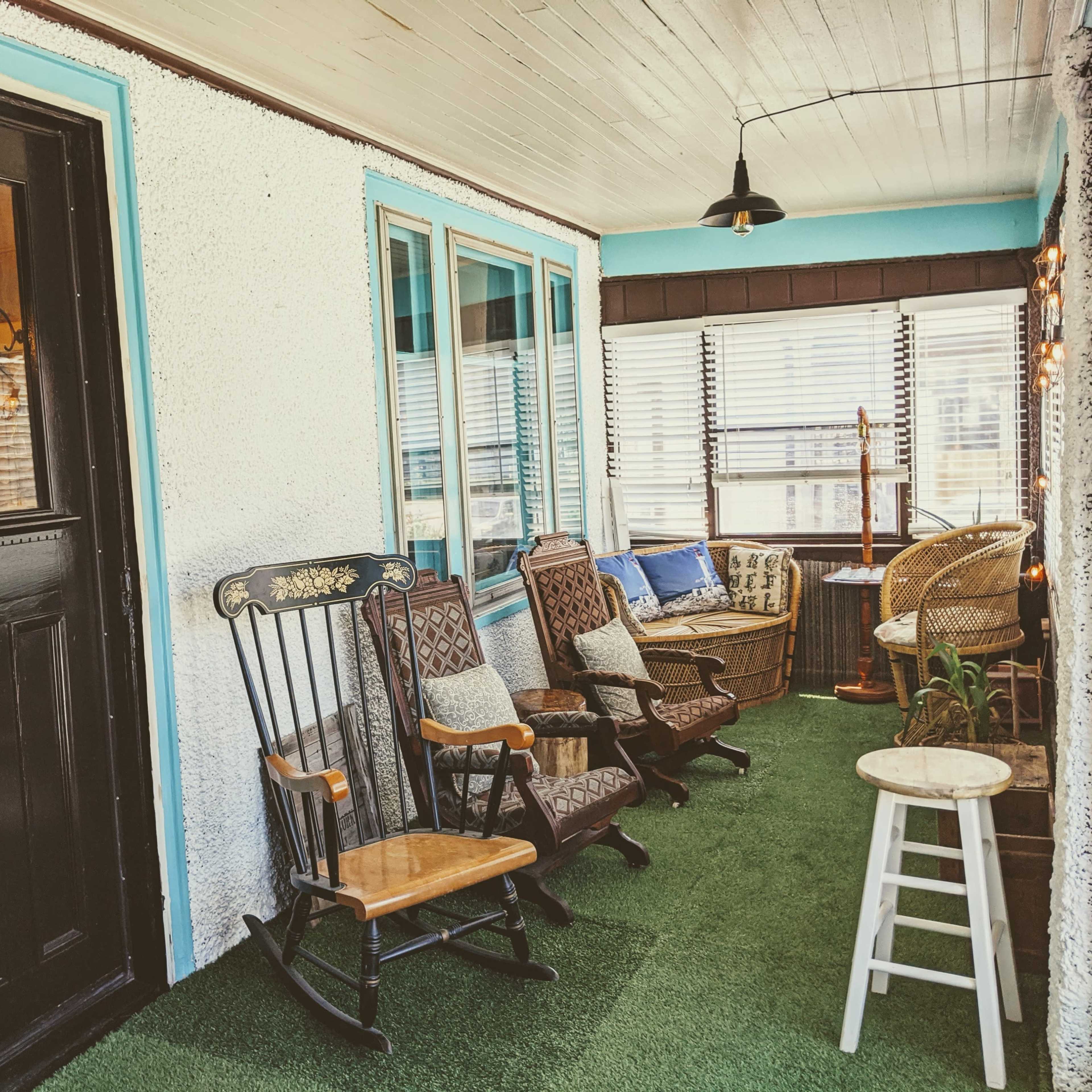 A bright, inviting porch furnished with several rocking chairs, a wicker chair, and a small side table, all set against a backdrop of large windows.