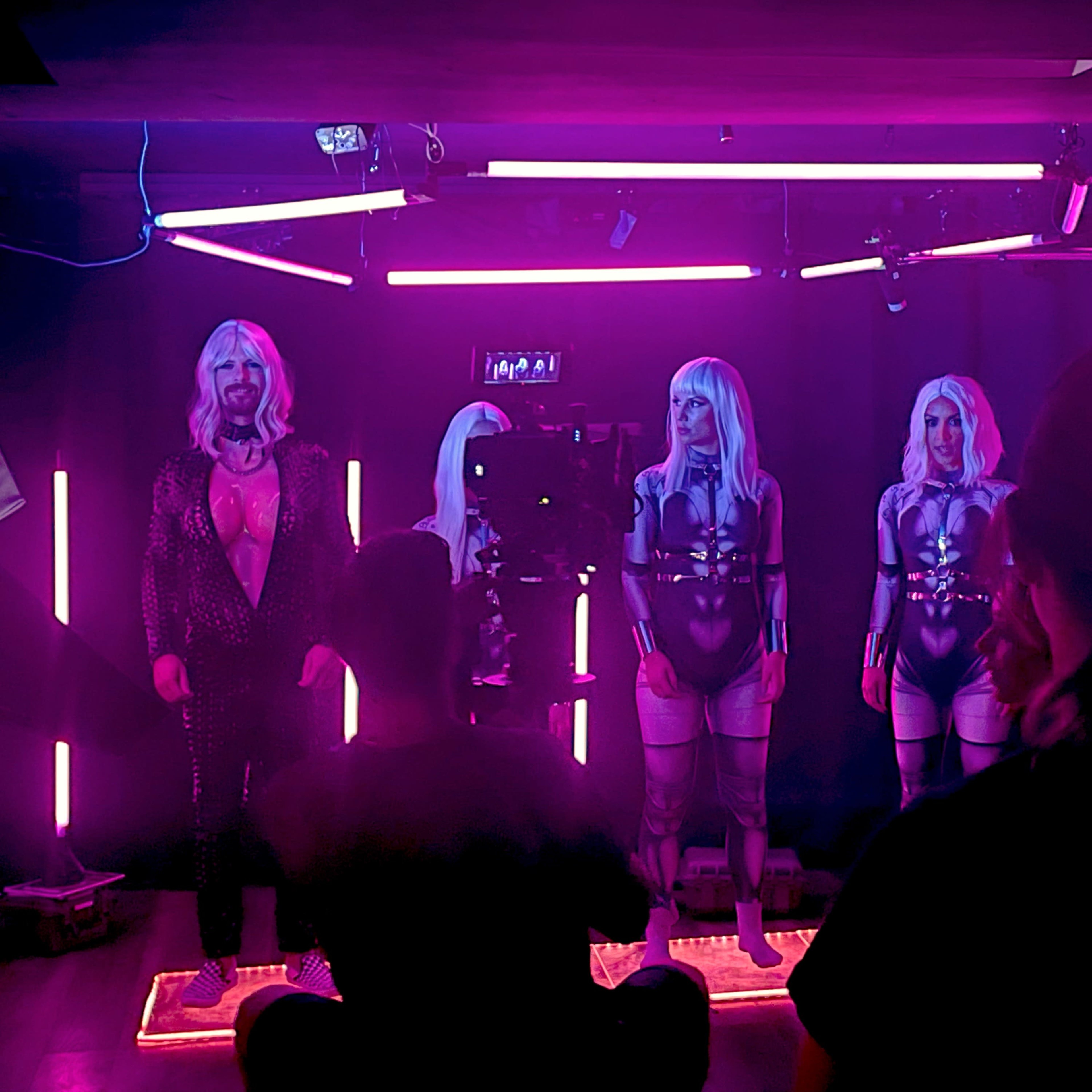 Three individuals with long hair and matching outfits pose in front of a camera in a room illuminated by pink neon lights.