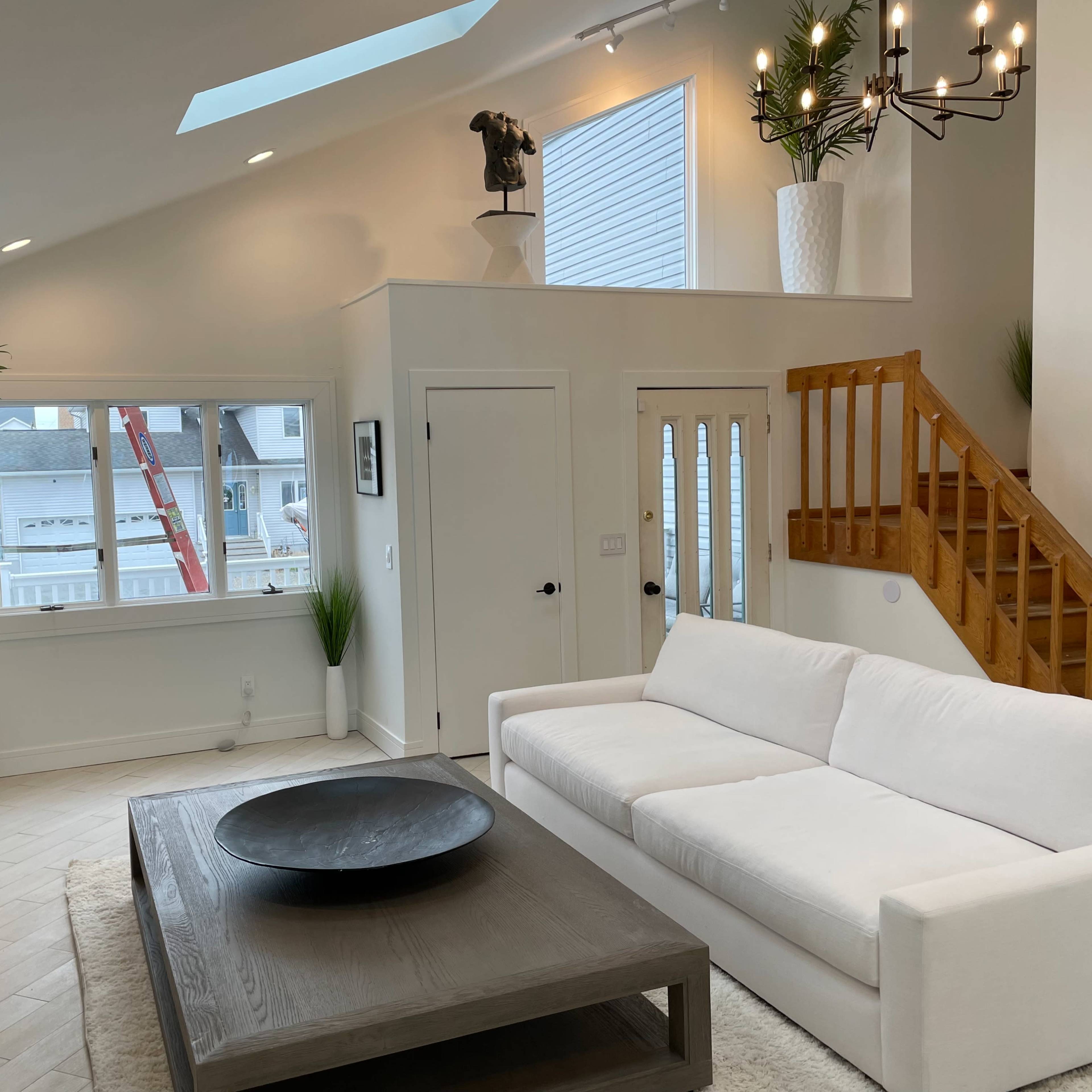 A modern living room featuring a white sofa, a round black coffee table, large windows, and a staircase leading to an upper level.