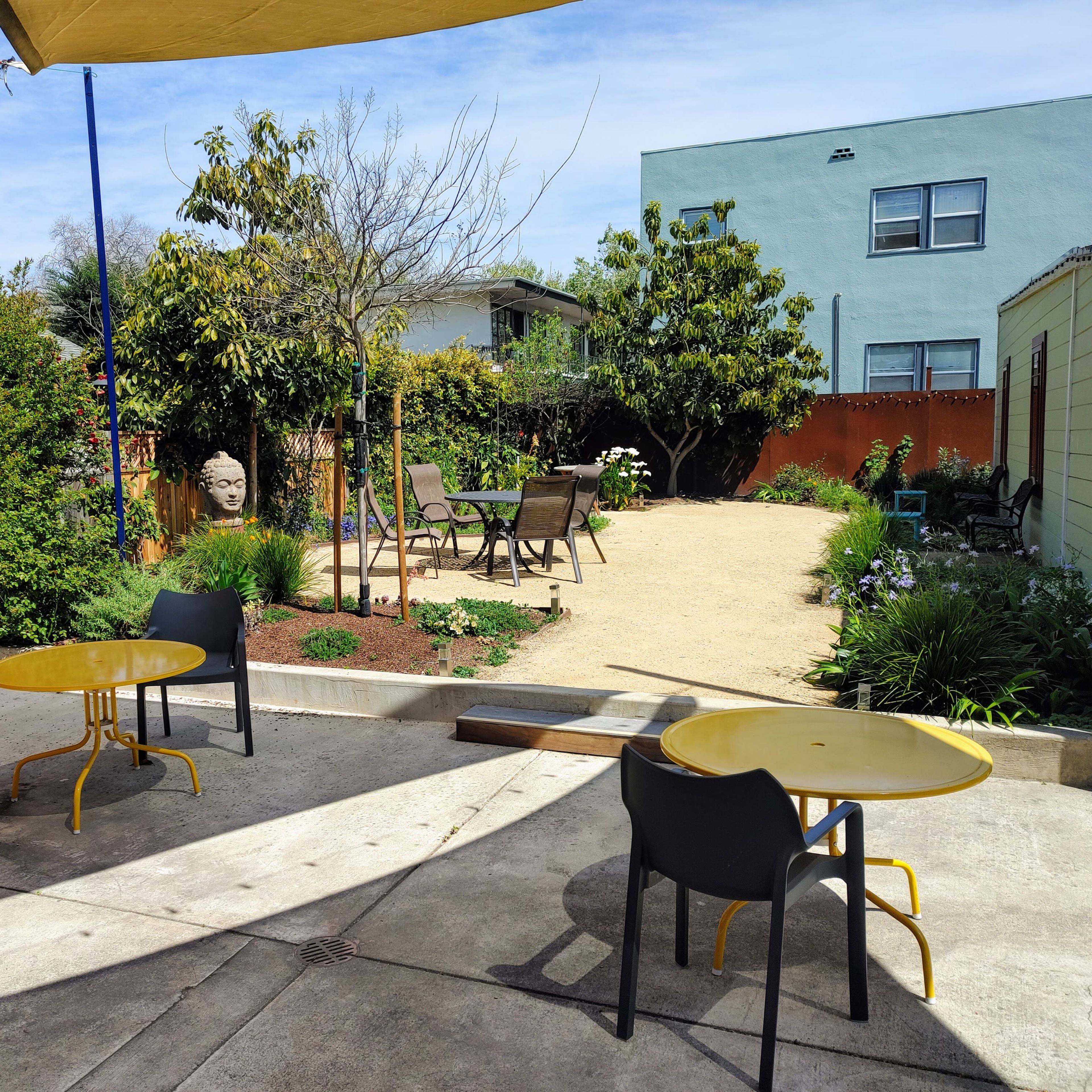 A backyard garden with two yellow tables and chairs in the foreground, surrounded by greenery and decorative elements.